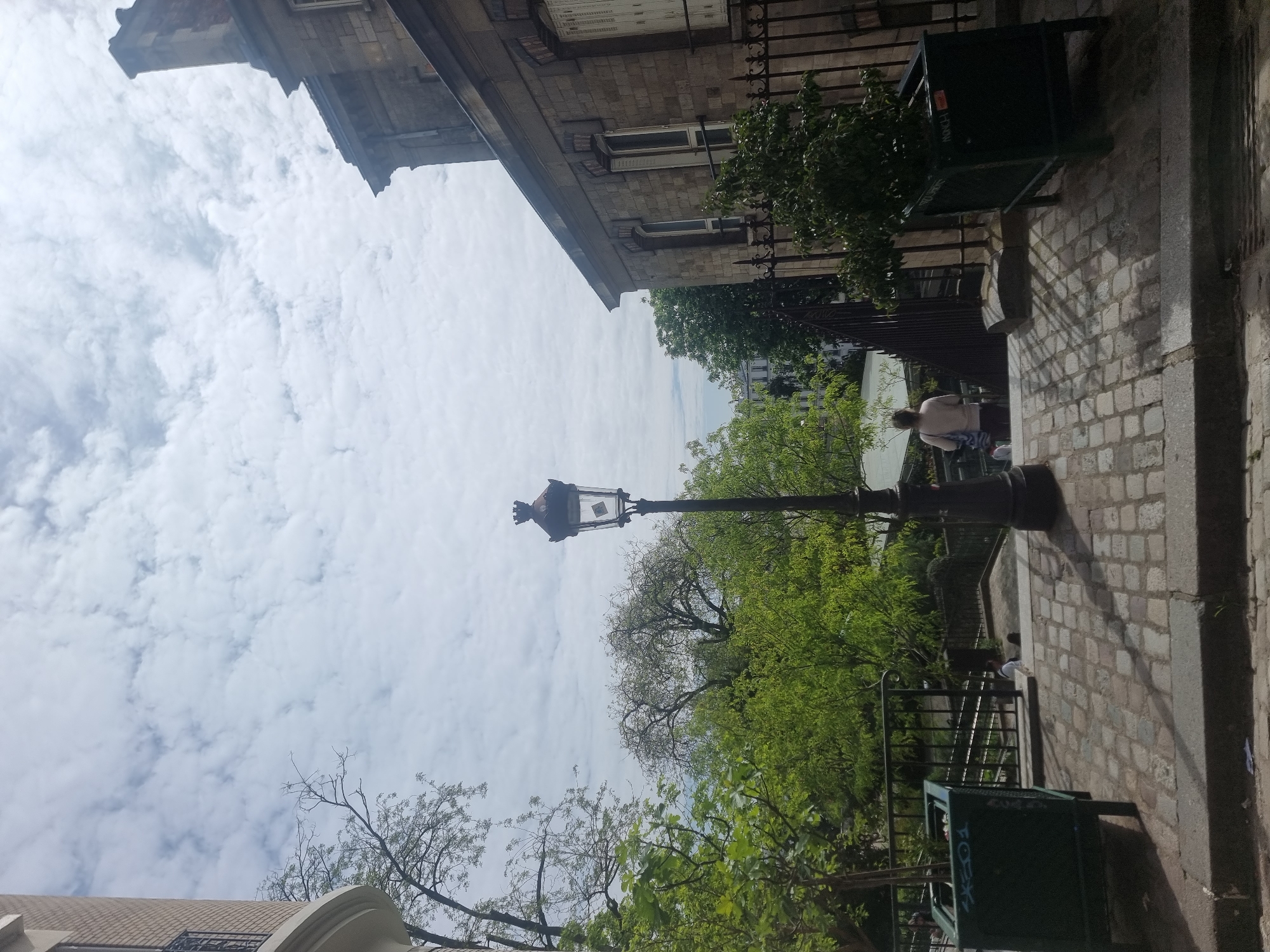 This photograph captures an outdoor urban scene in Paris, France, on an overcast yet bright day. The perspective is slightly elevated, looking down a pathway or street.
To the right, a classic Parisian building with a stone facade, ornate window frames, and several rectangular windows is prominent. A dark, decorative wrought-iron railing runs along its side. Another building's rooftop and upper section are visible in the upper left, contributing to the city's architectural character.
A tall, black, ornate street lamp with a traditional glass lantern design stands in the mid-ground. Lush green trees with dense foliage are abundant, framing the street lamp and extending into the background, with some bare branches visible in the foreground and distance, suggesting varied tree types or stages of leaf growth.
The ground on the right is paved with light-colored cobblestones, leading downwards and bordered by a concrete curb. Shadows are cast across the pavement, indicating the indirect light of an overcast day. In the lower right foreground, a dark green public bin is visible, featuring faint white graffiti that appears to spell out "FOX" and "NYC."
A person is seated on a low stone wall or bench in the mid-right portion of the image. They are wearing a light-colored top and dark trousers, with a striped bag or piece of fabric next to them. Another figure is faintly discernible further down the path. Black metal railings are present, separating different levels or areas of the scene.
The sky is filled with soft, scattered white and grey clouds, without direct sunlight. The overall impression is one of a calm, everyday street scene within an old European city.