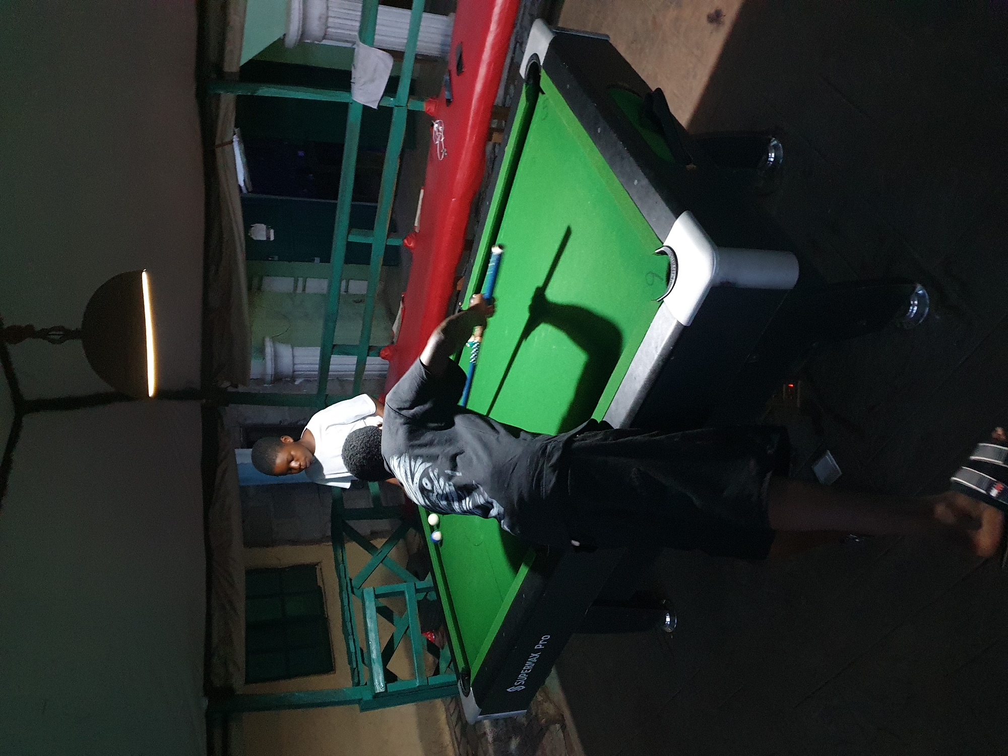 A man in a black shirt and shorts is playing pool on a Supermax Pro pool table with a blue cue stick. Another man in a white shirt is standing and watching. The pool table is set up on a patio with green railings. A red table is behind the pool table. The location is Lokoja, Nigeria.