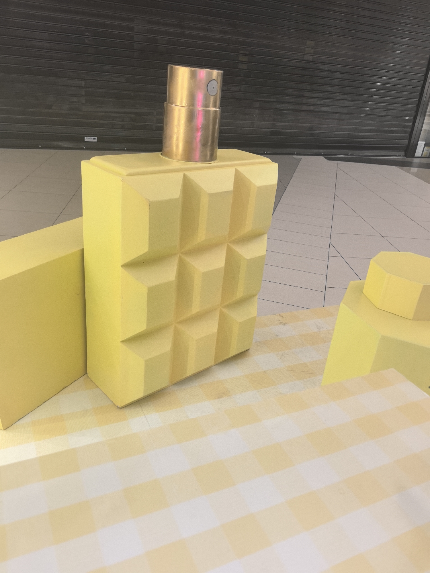 A large, whimsical, yellow display features oversized, geometric shapes resembling a perfume bottle and various other blocks, set against a background of a closed, gray rolling shutter. The “perfume bottle” has a gold-colored, cylindrical top. The display sits on a surface with a pale yellow and white checkered pattern. The location appears to be indoors, possibly in a shopping mall or commercial space, given the tiled floor and the presence of the closed shutter.