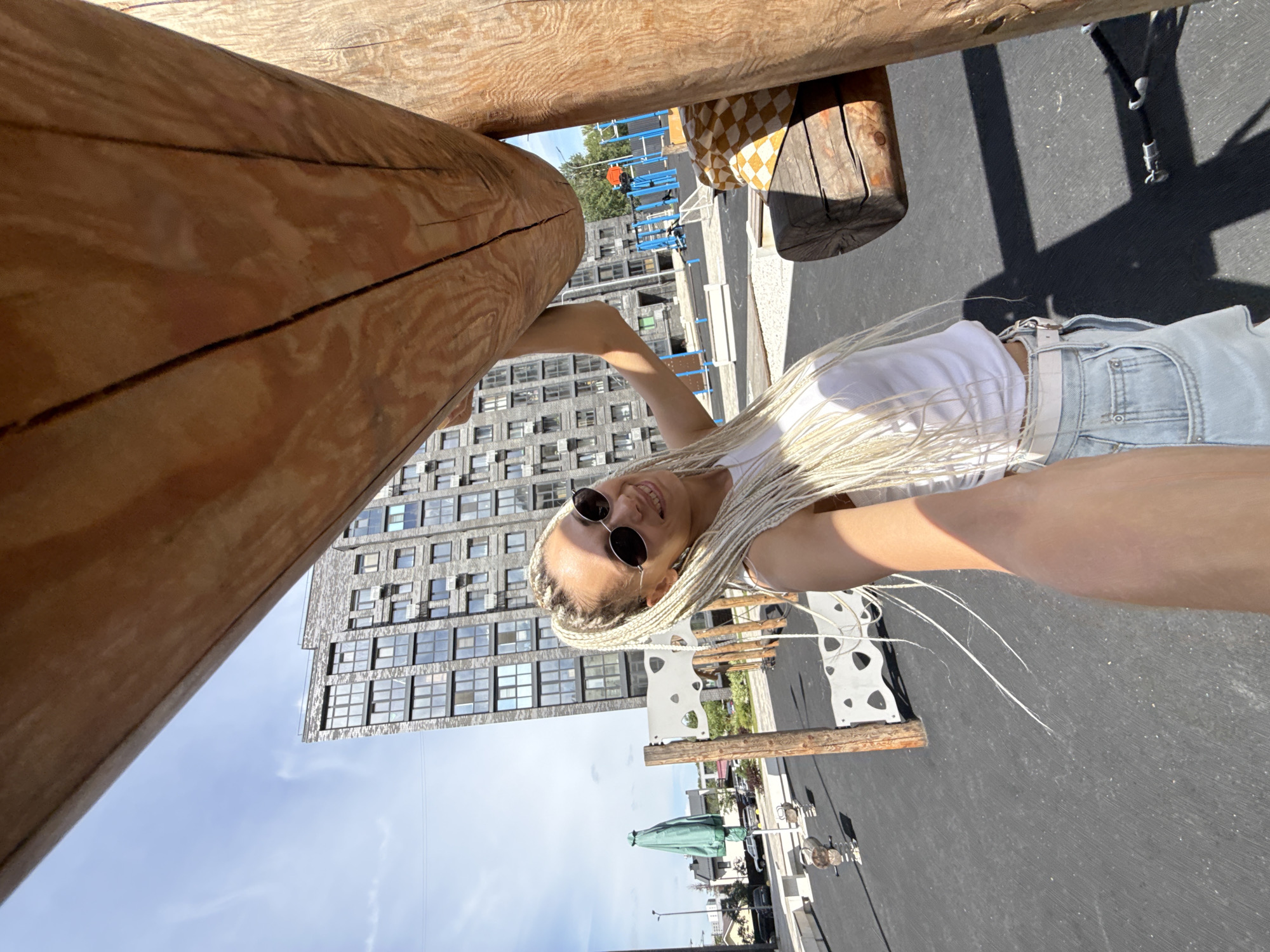 An outdoor, medium-wide shot captures a woman with long, blonde braided hair and sunglasses, wearing a white tank top and light-wash denim shorts. She is smiling and holding a large wooden beam, seemingly posing for a selfie. The background features a modern apartment building, a playground with blue structures, and a gray asphalt ground. The scene is well-lit by sunlight, casting shadows on the ground. The overall setting appears to be a contemporary urban residential area in Rusanovo, Russia.