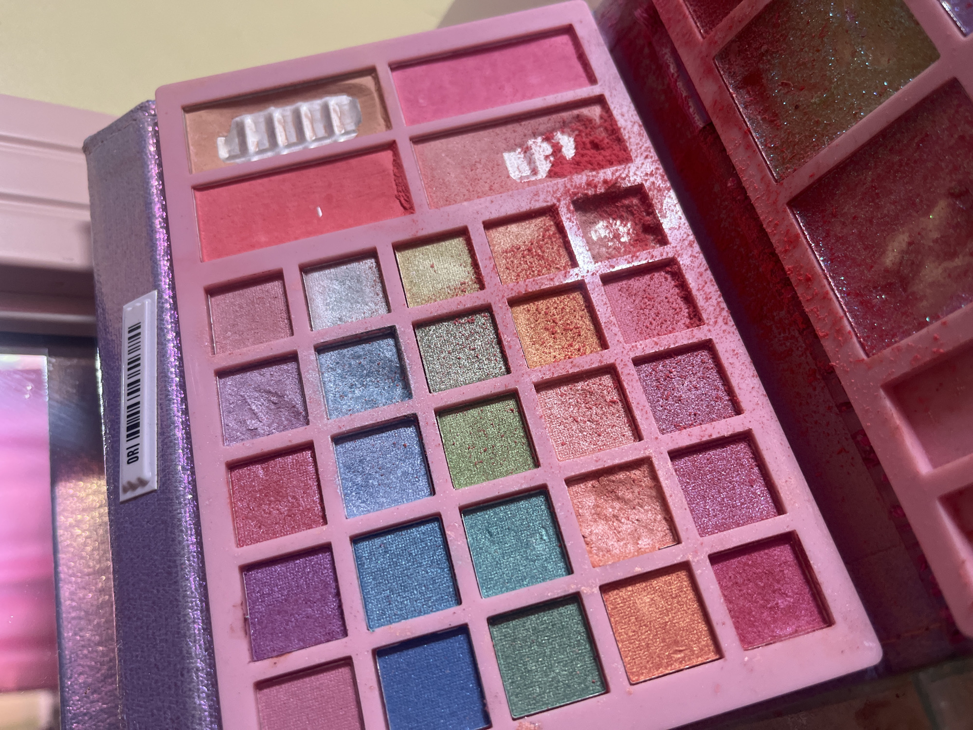 The image shows an open pink makeup palette with multiple eyeshadow and blush compartments. The palette is filled with various colors of pressed powder makeup, including purples, blues, greens, oranges, and pinks. Some of the compartments show signs of use, with powder spilled or smudged. The palette is positioned on a surface that appears to be a table or vanity. The background is blurred and shows a pink, reflective surface and a yellow wall. There is no indication of the city of Tarnivtsi, Ukraine, in the image.