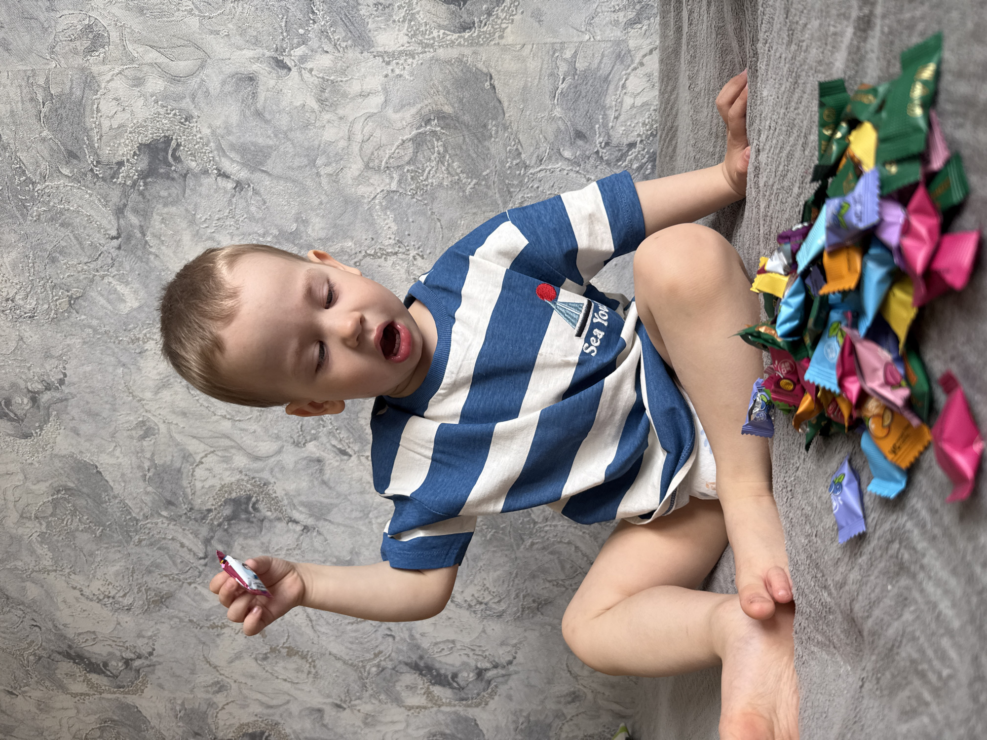 The image captures a young child, appearing to be a toddler or preschooler, sitting indoors in Rusanovo, Russia. The child has fair skin and light brown hair, wearing a blue and white striped short-sleeved t-shirt. The shirt features an embroidered graphic of a sailboat with a red circle and the partial text "Sea Yo...". A white diaper or underwear is visible beneath the shirt.
The child is positioned on a soft, grey textured surface, likely a blanket or couch. To their right, there is a large pile of individually wrapped candies in a vibrant assortment of colors, including green, purple, pink, yellow, blue, and orange. Some candy wrappers display Asian characters and English words like "blueberry." The child holds one small, colorful wrapped candy in their left hand. Their mouth is open wide, suggesting an expression of excitement, surprise, or anticipation, possibly about to eat the candy or reacting to the pile.
The background features a textured wall with a mottled grey and white decorative pattern. The scene depicts an activity centered around the child interacting with a large quantity of sweets, possibly after receiving them. The setting is a residential interior, likely during daytime.