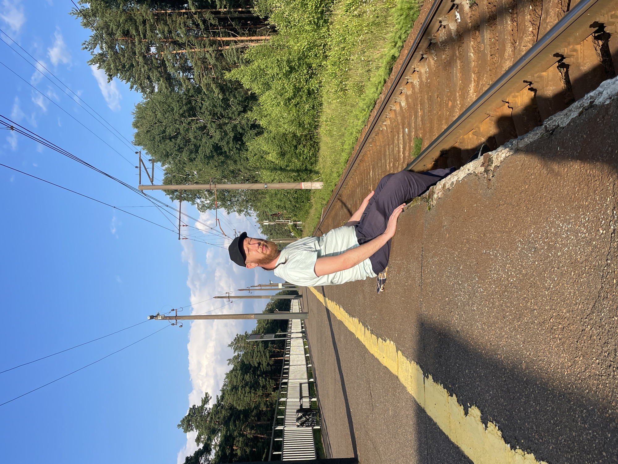 A man with a beard and a black baseball cap sits on the edge of a train platform in Lebyazhye, Russia. He is wearing a light blue t-shirt and dark blue pants. His right leg is bent at the knee, and his left leg is extended downwards. He is looking towards the camera with his eyes partially closed. A checkered blanket is placed on the ground beneath him. The platform has a yellow line running along its edge. To the right of the platform are train tracks, and beyond that, a grassy area with trees. The sky is blue with some white clouds. In the background, there are trees, power lines, and a white fence. A trash can and a bench are visible along the platform.