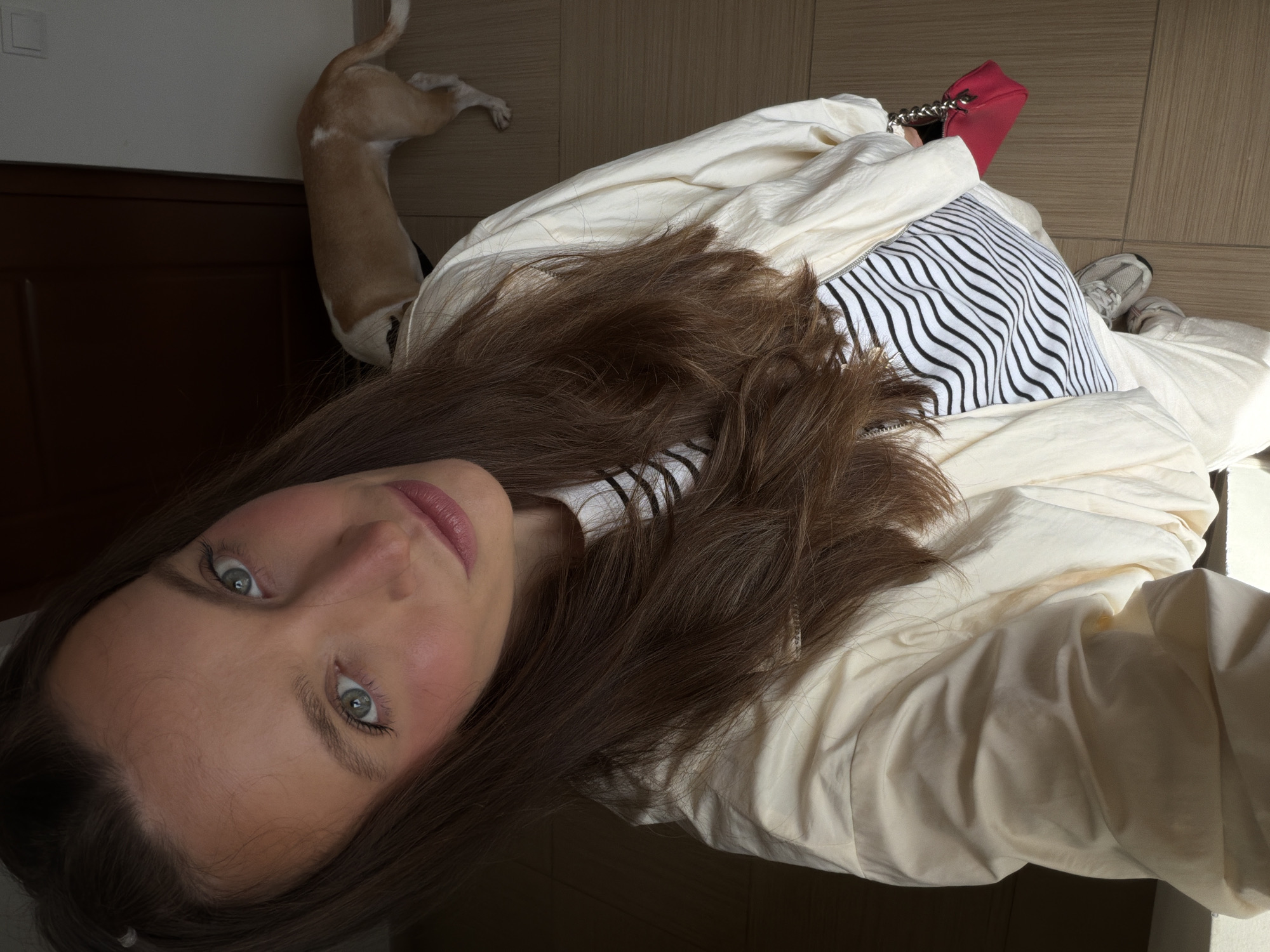 A woman with brown hair and light eyes is lying down, possibly on a bed or floor, in an interior setting. She is wearing a white shirt and a striped top. Beside her is a red purse and a pair of sneakers. In the background, a dog's hind legs and tail are visible. The location is possibly an interior in Budva, Montenegro.