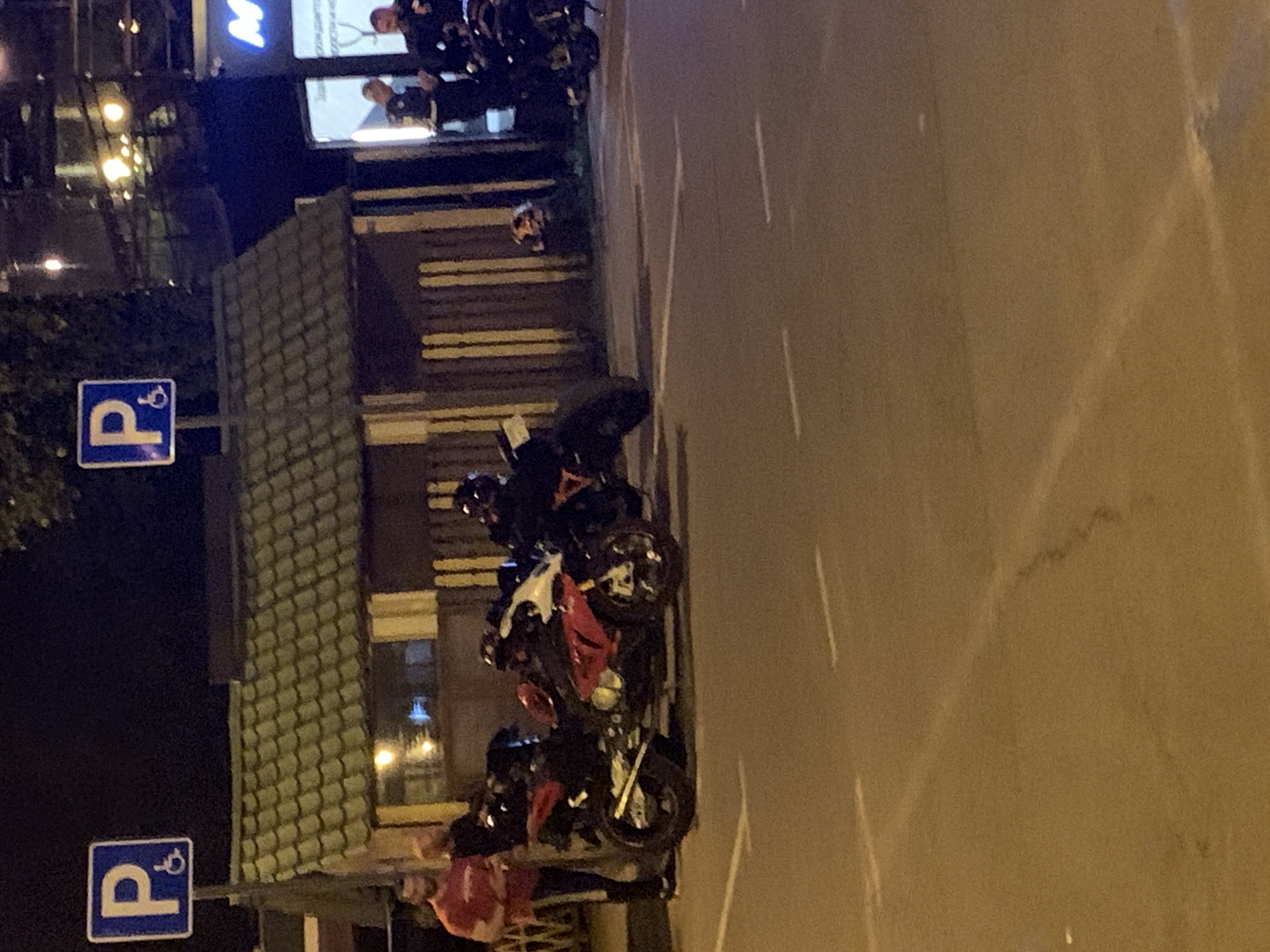 In an outdoor nighttime scene, several individuals and motorcycles are present. In the foreground, two motorcycles are parked side-by-side, with a third motorcycle partially visible to the right. One of the motorcycles in the foreground is red and white, while the other is black. Two men are standing near the motorcycles, one wearing a red t-shirt and the other in dark clothing. Further in the background, several more individuals are standing near a building with a green tiled roof. One person is wearing a white shirt and black pants, and another is wearing a dark uniform. A blue and white sign indicating parking and disabled access is visible in the upper left corner. The ground appears to be asphalt with painted white lines, suggesting a parking area. The overall lighting is dim, characteristic of nighttime.