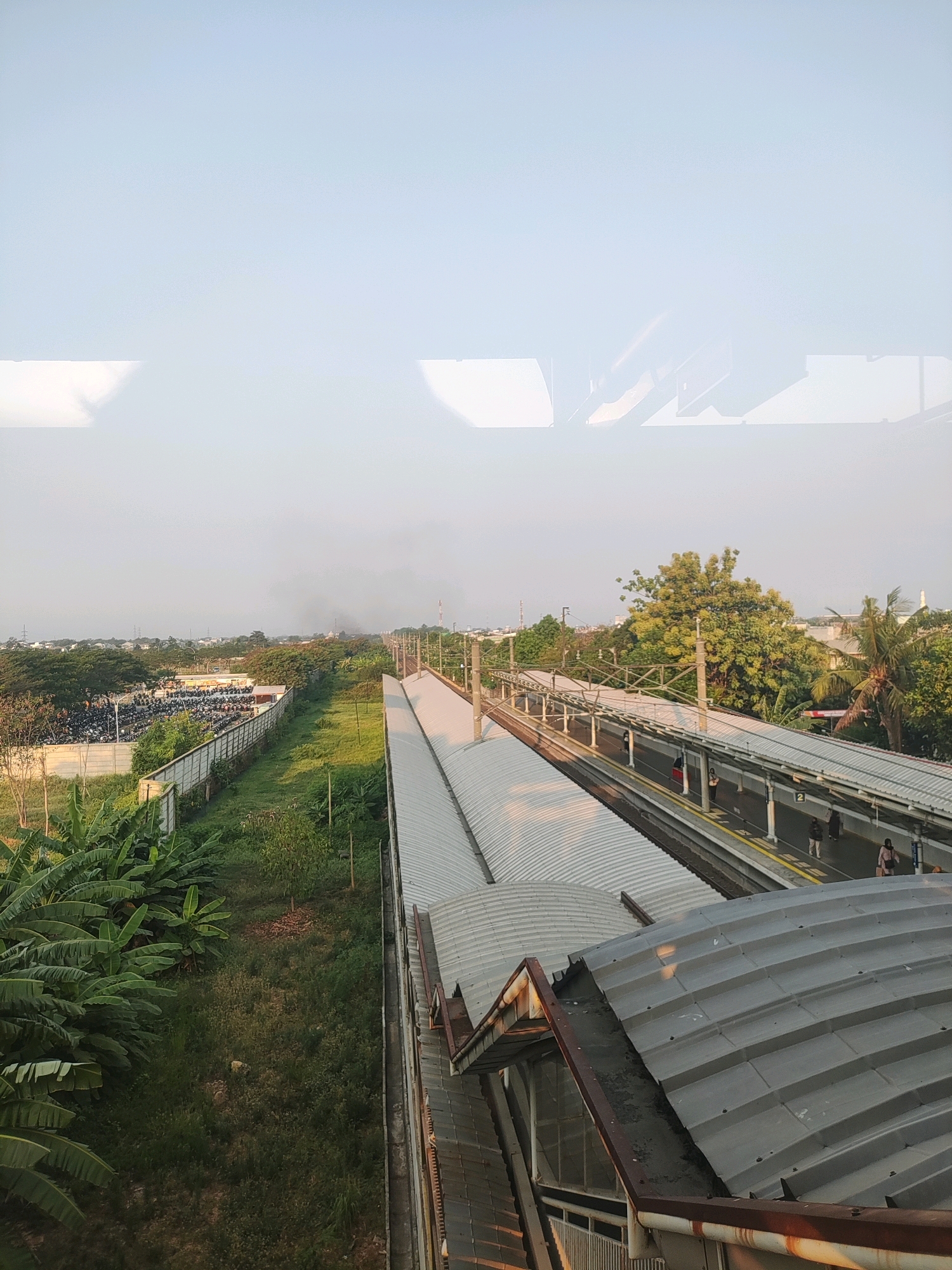 The image depicts an outdoor scene at a train station, likely in Telagamurni, Indonesia, given the tropical vegetation and architecture. In the foreground, a corrugated metal roof of a building or walkway is visible, showing signs of rust. Beyond this, a train track runs parallel to a station platform with a white canopy overhead. Several individuals are present on the platform, with some standing and others walking. The platform is bordered by yellow lines and has signage indicating platform numbers.
To the left of the tracks, a green, grassy area is interspersed with banana trees. Further to the left, a fence separates the grassy area from a large parking lot filled with motorcycles. In the distance, a hazy sky suggests either early morning or late afternoon lighting, with some industrial-looking structures and trees dotting the landscape. Electrical poles and wires are also visible, indicating the presence of infrastructure. The overall impression is of a functional, active train station within a semi-urban or peri-urban environment.