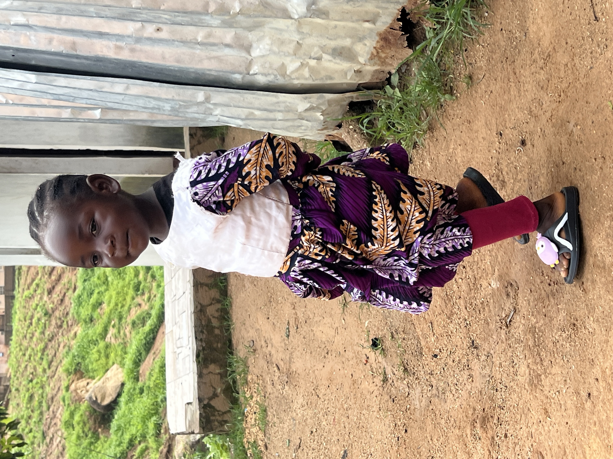 A young Black girl, likely under the age of seven, stands on a dirt path in Jos, Nigeria. She is wearing a white top with puffed sleeves that transitions into a vibrant purple and orange patterned dress. She has on bright pink leggings and black sandals with a pink cartoon character on the left strap. The girl is facing the camera, her head turned slightly to her left. Her hair is styled in braids. Behind her, a corrugated metal structure is visible to the right, with overgrown grass at its base. To the left, a grassy embankment with a large rock is present, along with a concrete bench-like structure. The ground is uneven and dusty.