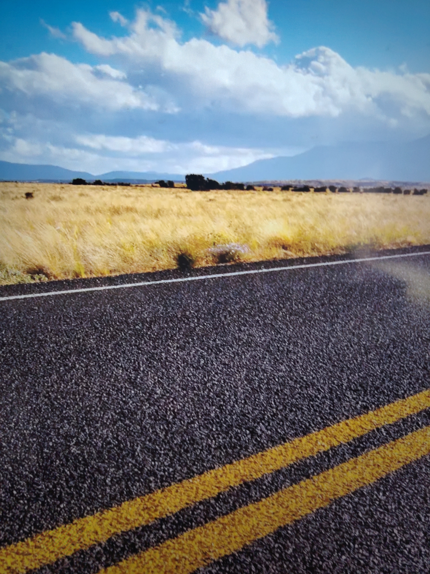 A low-angle, ground-level shot presents a rural scene under a sky filled with clouds. The foreground is dominated by a paved road with double yellow lines indicating no-passing. The asphalt texture is visible, and a single white line separates the road from the adjacent landscape. This landscape features dry, golden grass extending to the horizon. The background reveals distant, hazy mountains. The location is unknown, but the scene suggests a rural environment. No people are visible.
