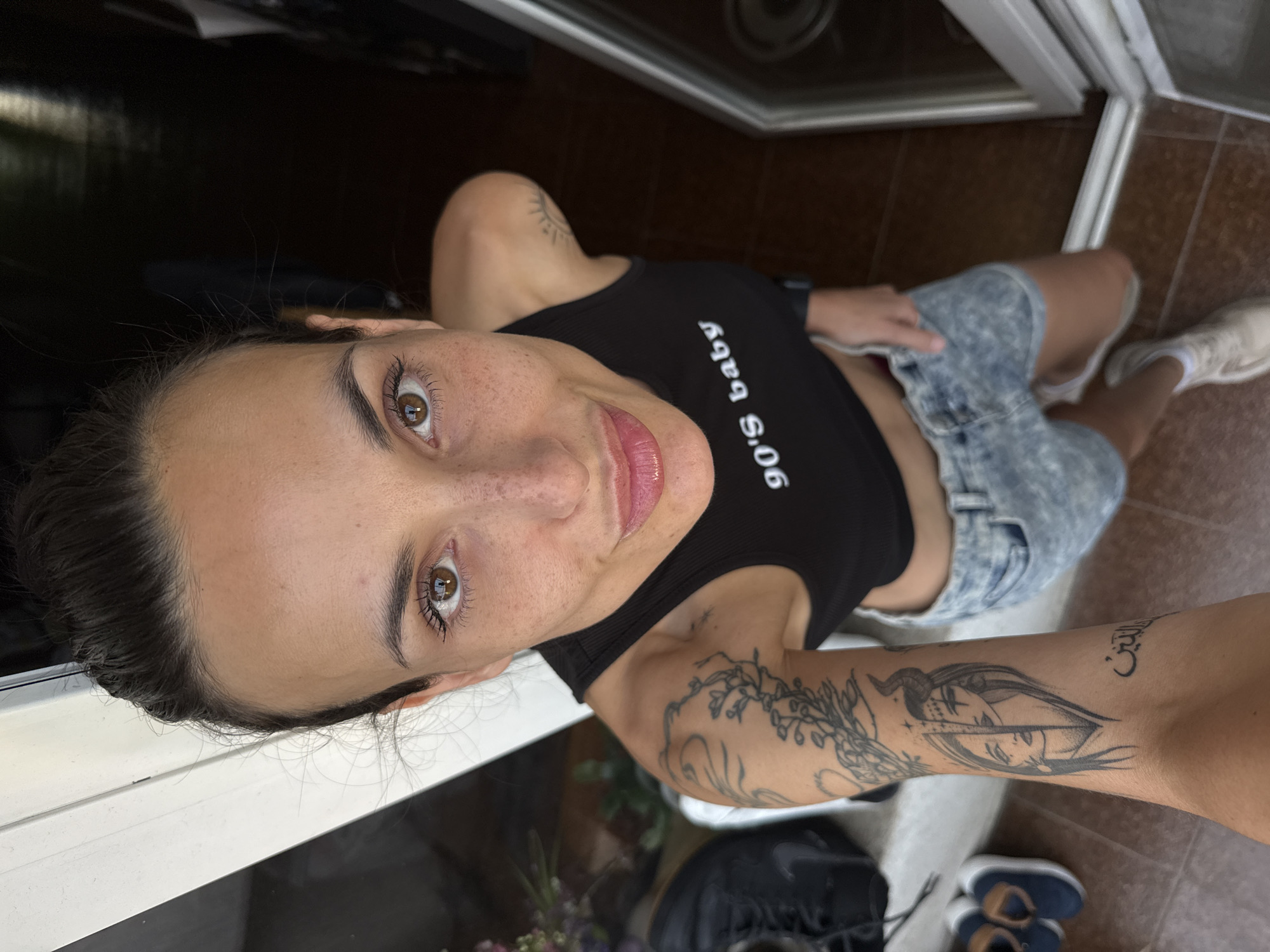 A woman with brown eyes and dark hair is taking a selfie from a high angle, looking directly at the camera. She has freckles and is wearing a black tank top with the text "90's baby" printed on it, along with acid-wash denim shorts. Tattoos are visible on her arms, including one of a female figure with horns. She is standing on brown tiled flooring near a white framed window or doorway. The location in the city of Pleven, Bulgaria is not identifiable in this image. The scene includes shoes and other items on the floor.