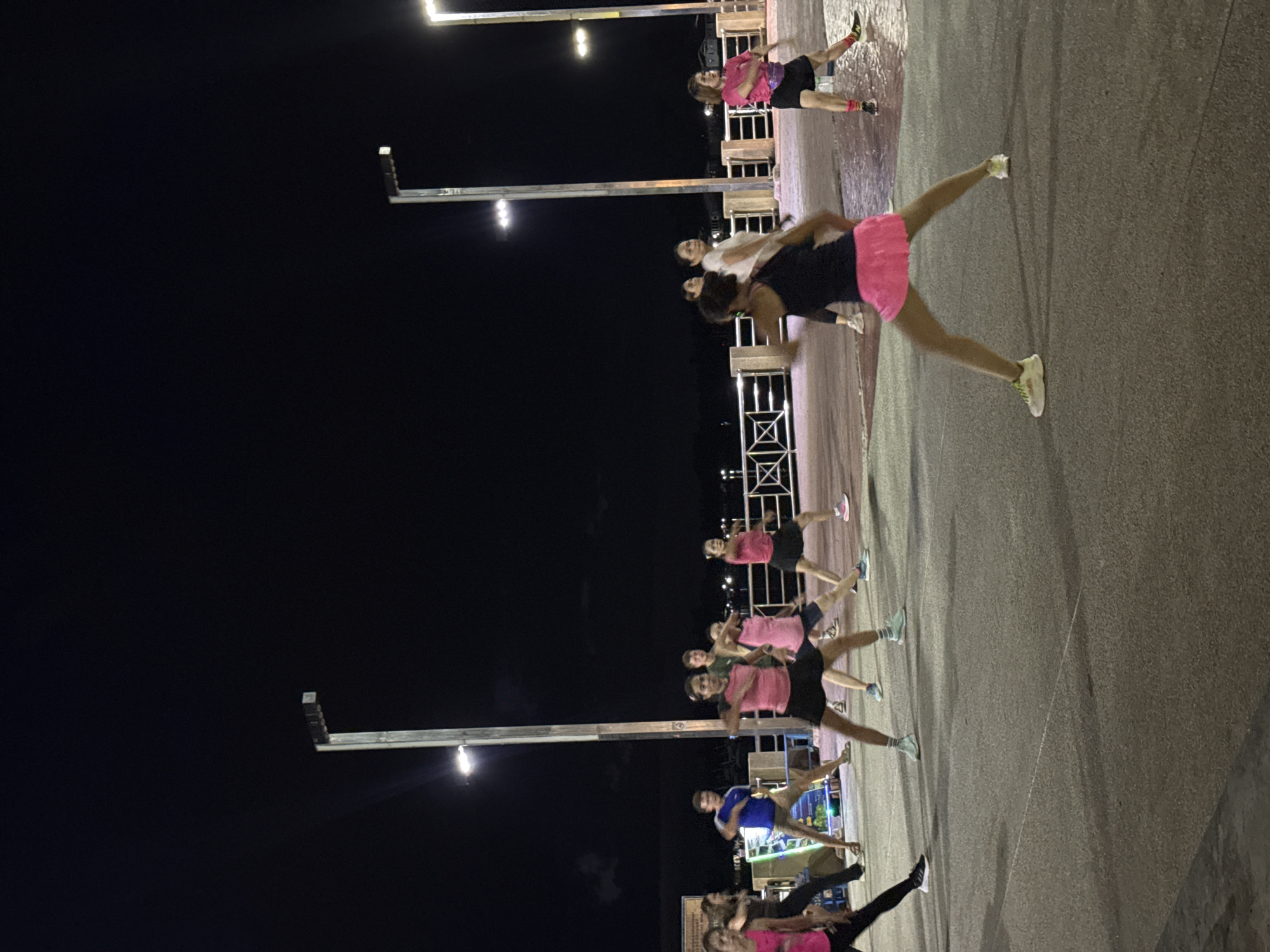 This image depicts an outdoor fitness class taking place at night on a paved walkway near a body of water. The location is identified as Surat Thani City Municipality, Thailand. Several individuals, predominantly women, are participating in a group exercise, demonstrating synchronized movements such as arm extensions and leg stances. They are dressed in athletic wear, including shorts, skirts, and t-shirts, in various colors like pink, black, and blue. The environment is illuminated by streetlights, creating distinct shadows on the ground. A metal railing separates the walkway from the water, and in the background, structures and lights suggest urban development. The overall scene conveys a sense of community and active recreation in a public urban space.