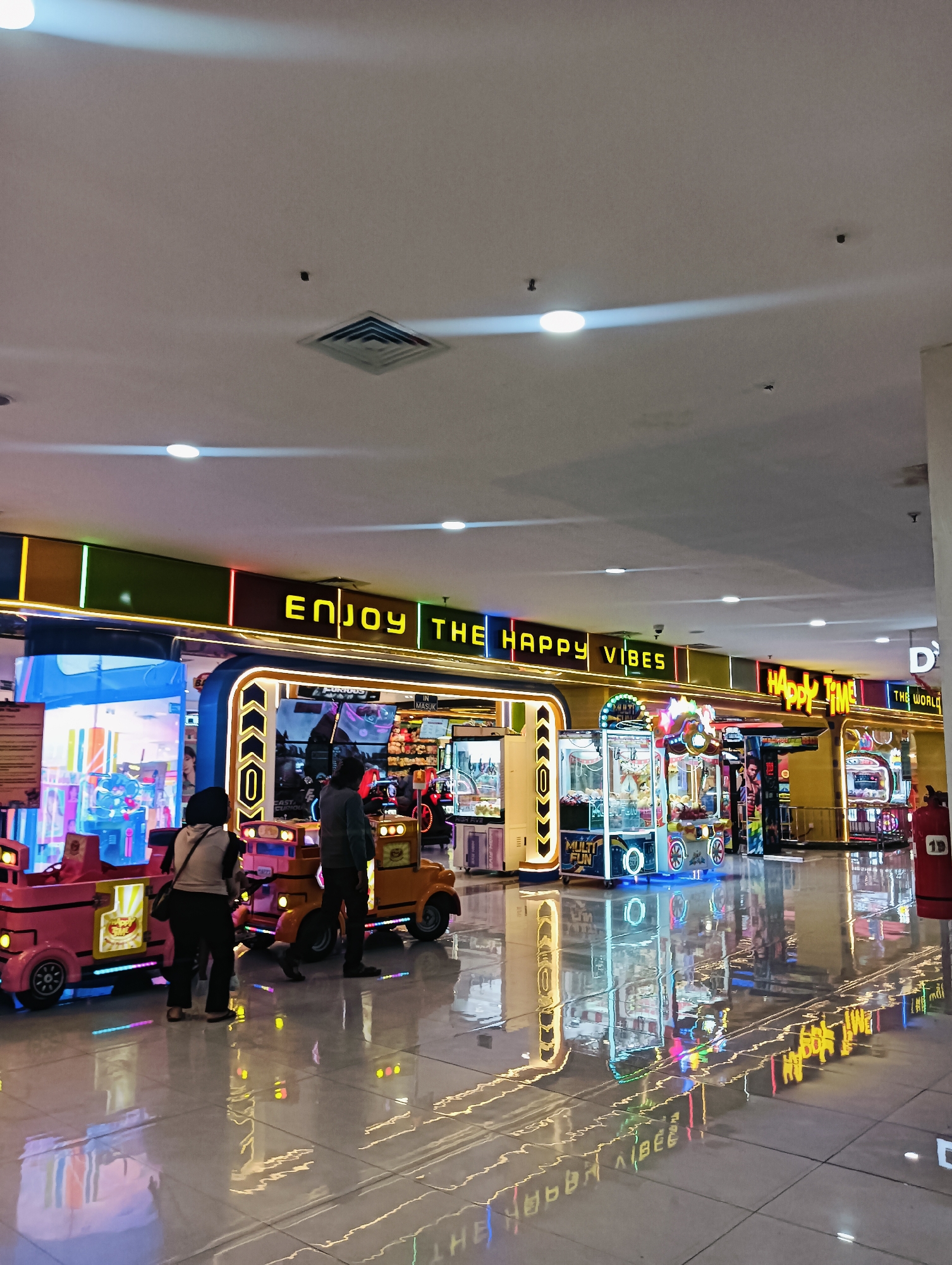 The image depicts the interior of an arcade located in Batam, Indonesia. Two individuals, a woman and a man, are standing next to brightly lit arcade game machines and kiddie rides. The arcade is characterized by neon signage that reads "ENJOY THE HAPPY VIBES" and "HAPPY TIME". The floor is highly reflective, mirroring the colorful lights and signage, creating a vibrant and dynamic atmosphere. Several arcade game cabinets, including claw machines, are visible, stocked with plush toys. The overall scene suggests a lively entertainment venue, likely a popular spot for families and individuals seeking recreation.