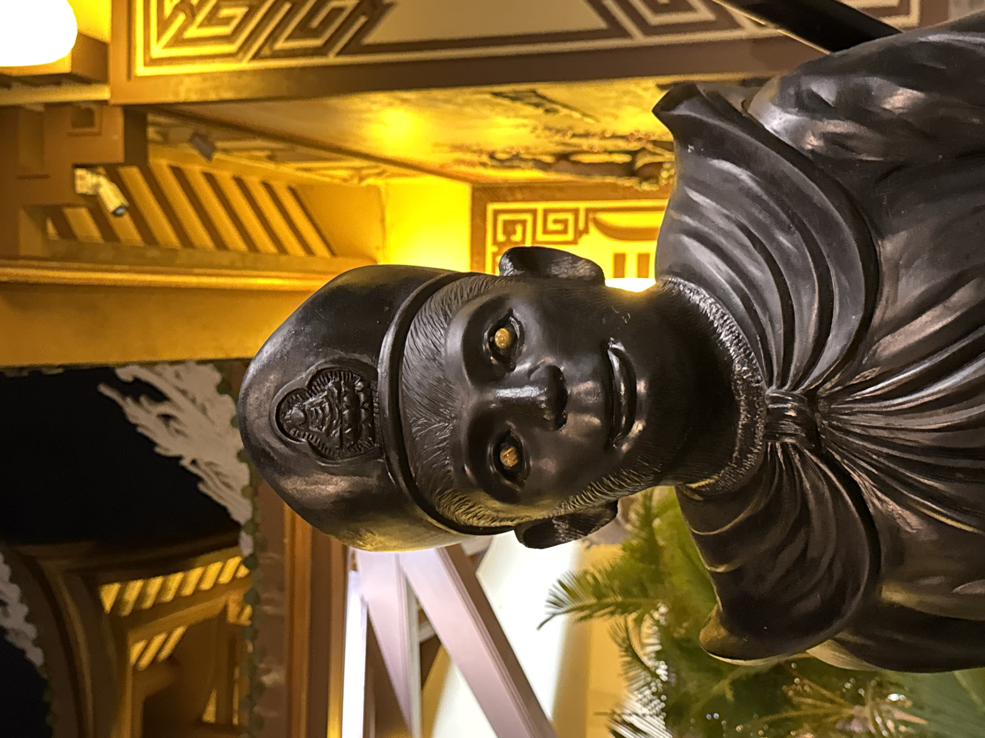 A close-up, low-angle shot shows a dark, weathered bronze statue of a monkey wearing a conical hat. The statue's eyes are a bright, reflective gold, and its mouth is open in a slight smile. The statue appears to be located in a temple or shrine, with warm yellow light illuminating the background. In the distance, elements of ornate architecture with intricate patterns and hanging decorations are visible, along with glimpses of lush green foliage. The overall impression is one of solemnity and reverence, possibly associated with Buddhist traditions, as suggested by the Buddha imagery on the statue's hat. This statue is likely located at the Linh Ung Pagoda on Son Tra Peninsula, Da Nang, Vietnam, a well-known tourist destination featuring a large statue of the Goddess of Mercy and other religious effigies.