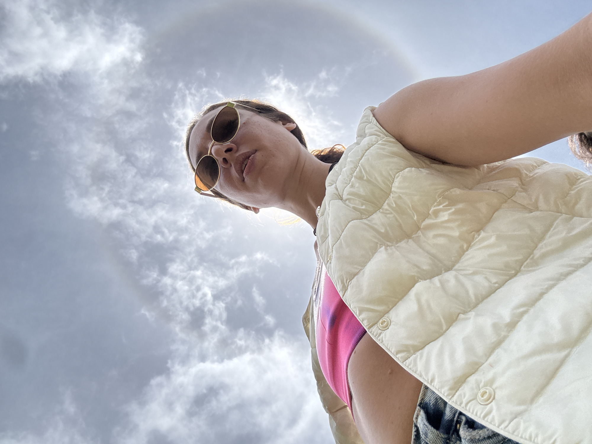 A low-angle, upward shot captures a woman from the chest up against a cloudy sky, which features a prominent sun halo. The woman, wearing sunglasses and a pink top layered with a cream-colored puffer vest, looks towards the camera. Her left arm is bent and resting on the vest. Jeans are visible at the bottom of the frame. The sun halo, a circular optical phenomenon, is clearly visible around the sun, creating a diffused light. The overall impression is an outdoor setting, possibly a scenic overlook, given the dramatic sky. The location is identified as Ta Xua, Vietnam.