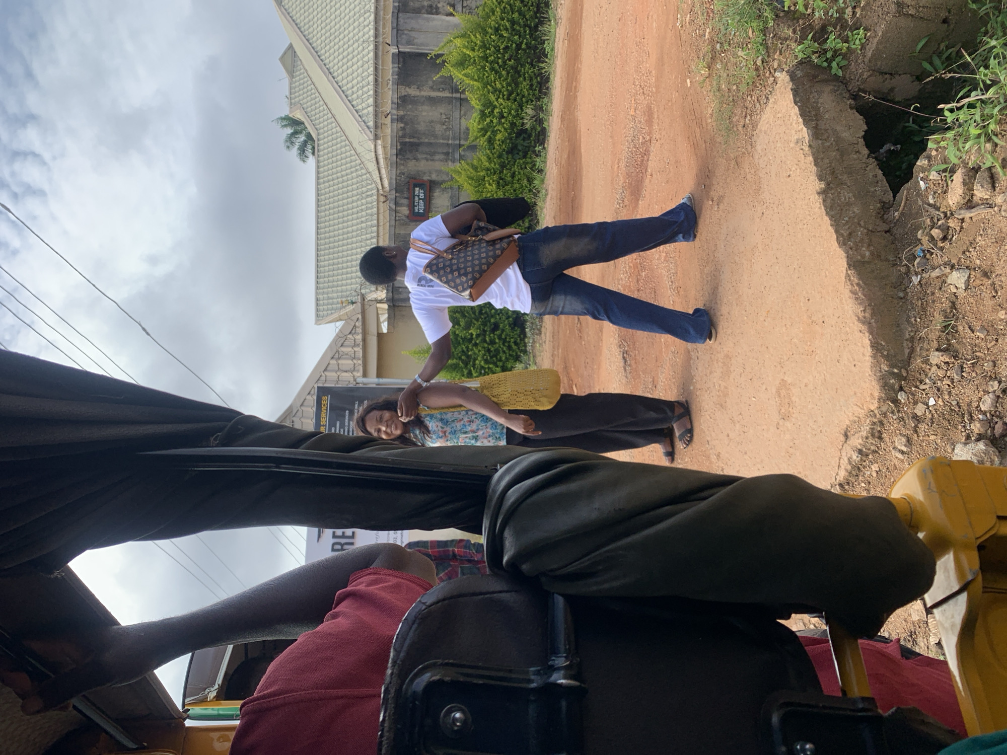 In Gura Top, Nigeria, a scene unfolds on a dusty, unpaved road. A woman, wearing a floral print blouse and dark pants, stands near a building with a sign reading "UR SERVICES." She smiles broadly, her arm extended as if being greeted or signaled. Beside her, another woman in a white t-shirt and blue jeans walks away from the camera, carrying a patterned handbag and a yellow woven bag. She appears to be leaving the area, her back to the viewer. The environment is characterized by red earth, sparse vegetation, and a cloudy sky overhead. The presence of a broken concrete curb and an open drainage ditch suggests an unmaintained urban or semi-urban area. In the foreground, part of a yellow motorized tricycle is visible, hinting at the mode of transportation in this locale. A person in a red shirt is seated inside the tricycle, partially obscuring the view. The overall impression is of an everyday street scene in Nigeria, with human interaction set against a backdrop of typical urban infrastructure and natural elements.