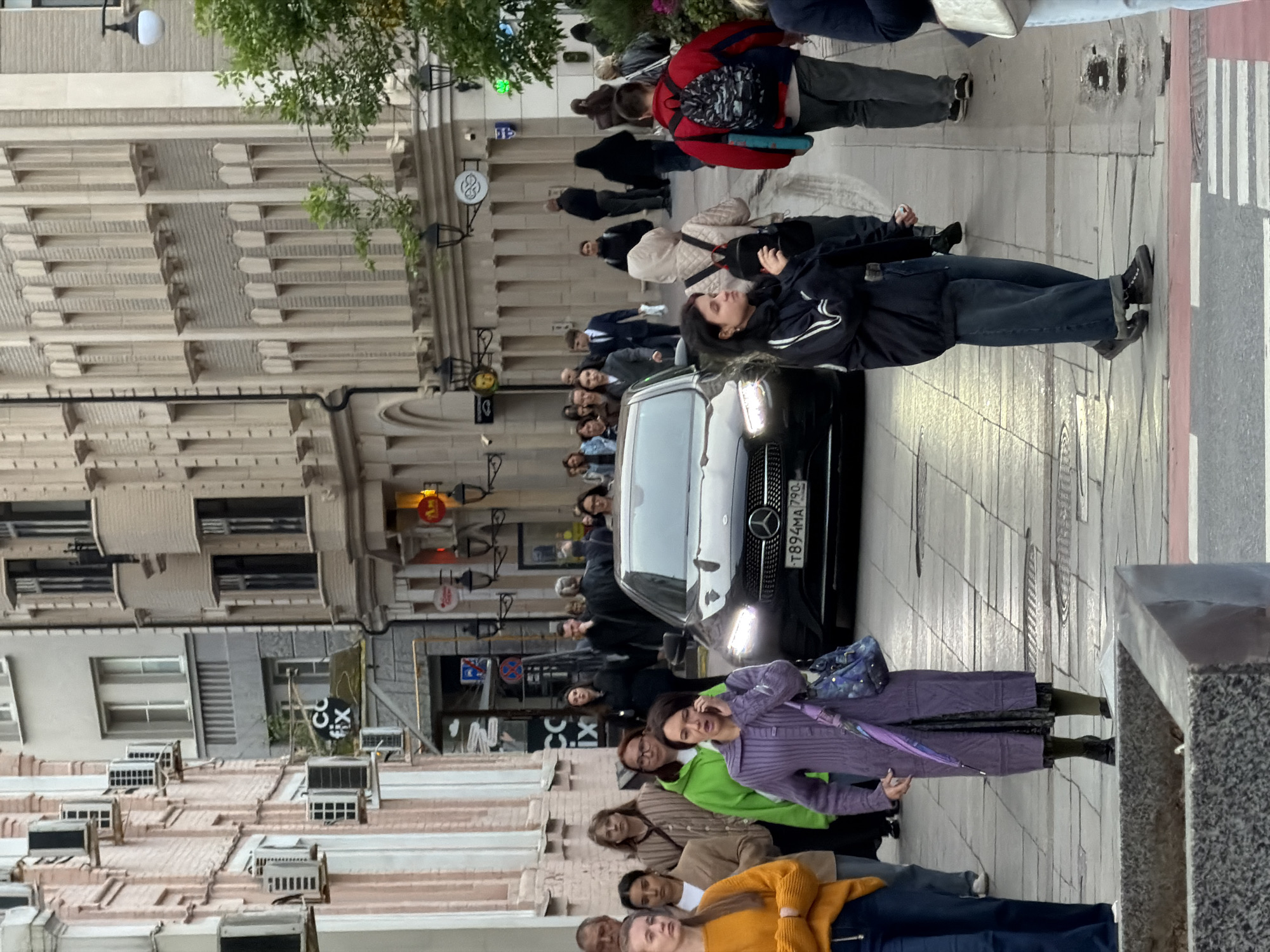 The image shows a street scene in Moscow, Russia. Pedestrians are walking on the sidewalk and crossing the street. A black Mercedes-Benz car with a license plate that reads "T894MA 790" is stopped in the street, possibly waiting for pedestrians to cross. The buildings lining the street are varied in architectural style. A sign with the letters "CO FIX" is visible.