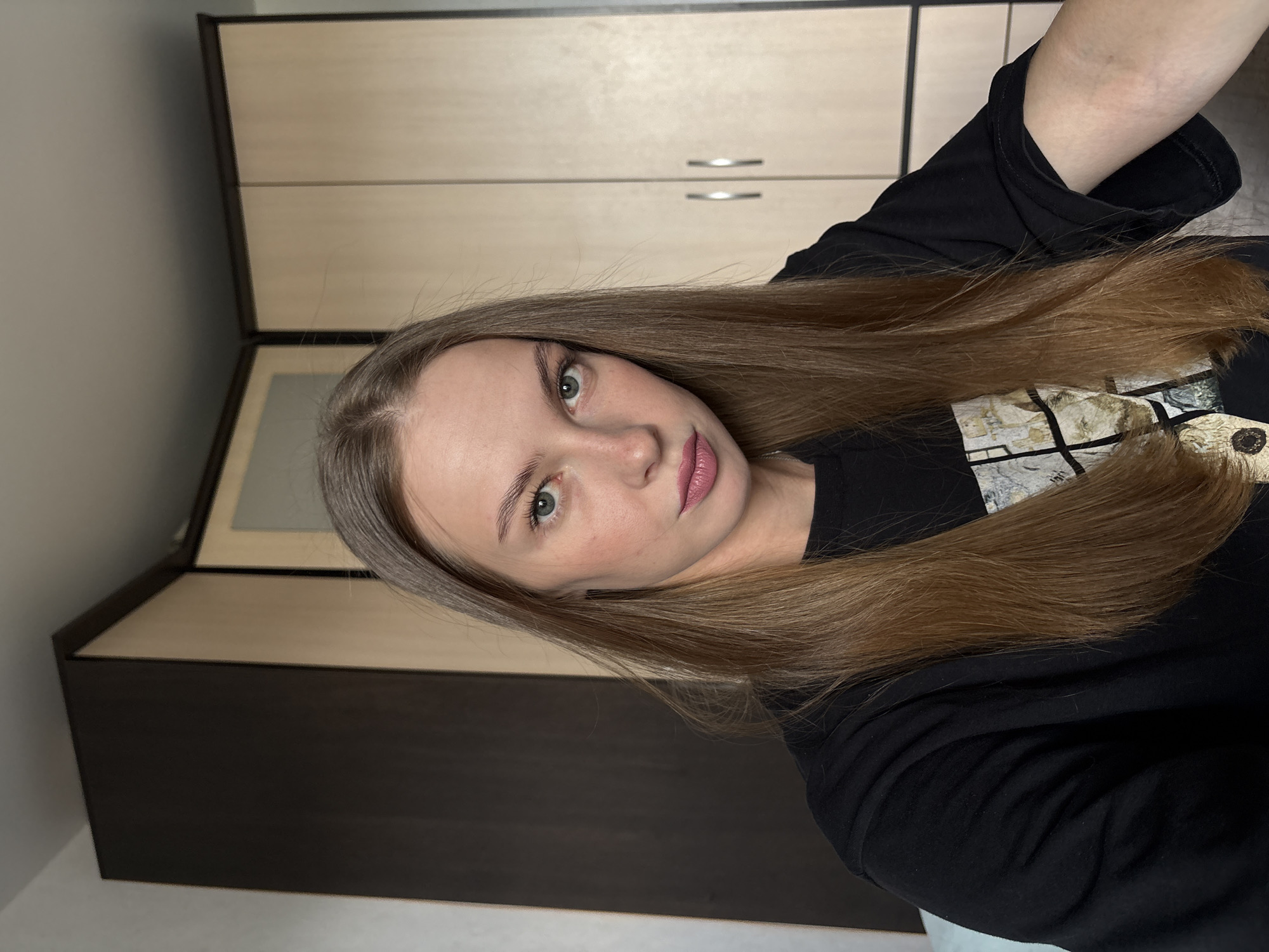 The image shows a young woman with shoulder-length brown hair and green eyes. She is wearing a black t-shirt and is looking at the camera with a neutral expression. The background appears to be a room with light-colored walls and a wooden wardrobe with dark brown doors. The overall lighting is soft and natural. The image was taken in Perm, Russia.