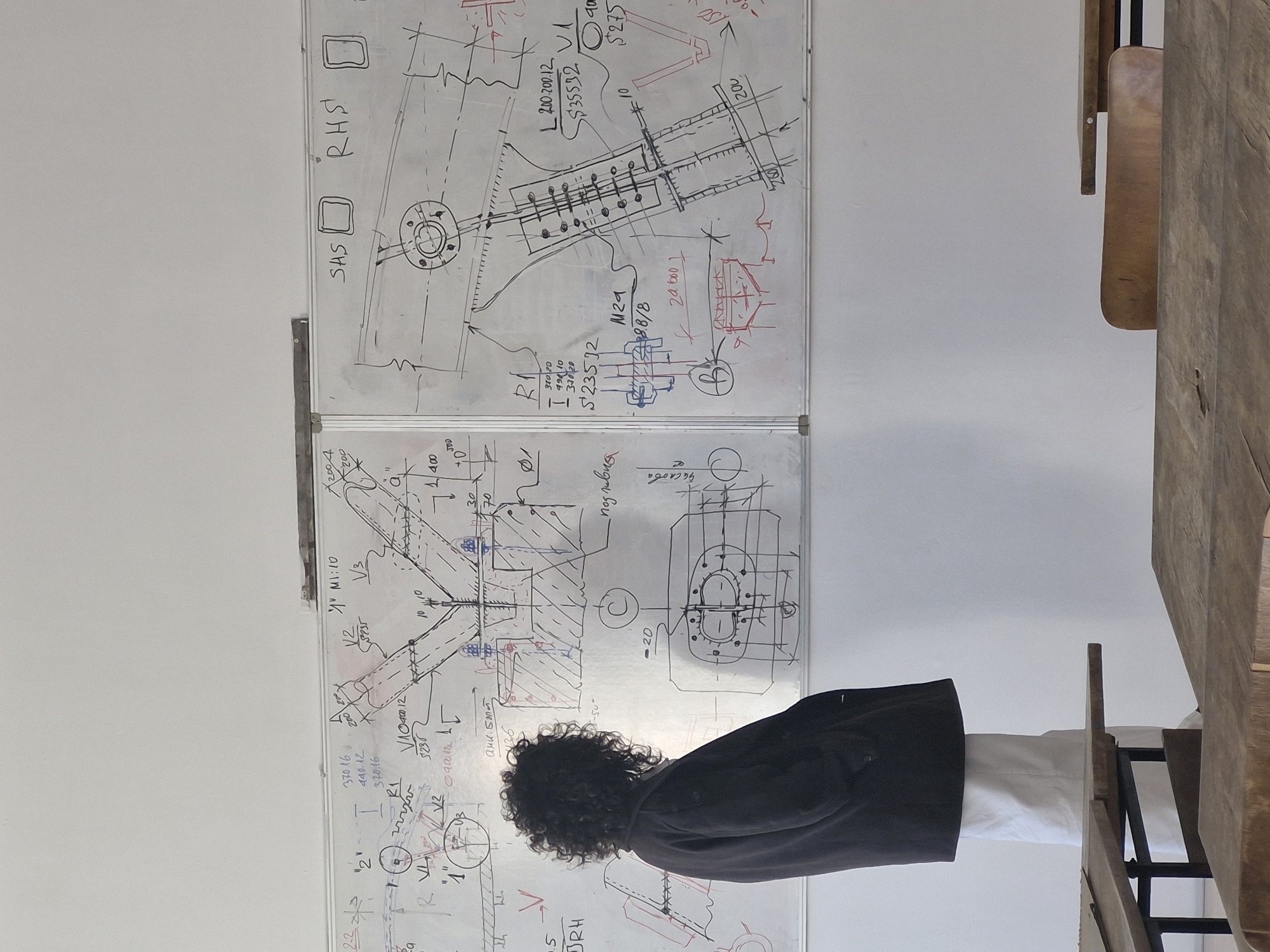 The image shows a person with dark, curly hair standing in front of a wall covered in technical drawings on whiteboards. The person is wearing a black jacket and a white shirt. The drawings appear to be engineering or architectural schematics, with annotations and dimensions written in black ink. A wooden table and chair are visible to the right of the person.
There is no information available to determine the location, let alone the city of Sofia, Bulgaria.
