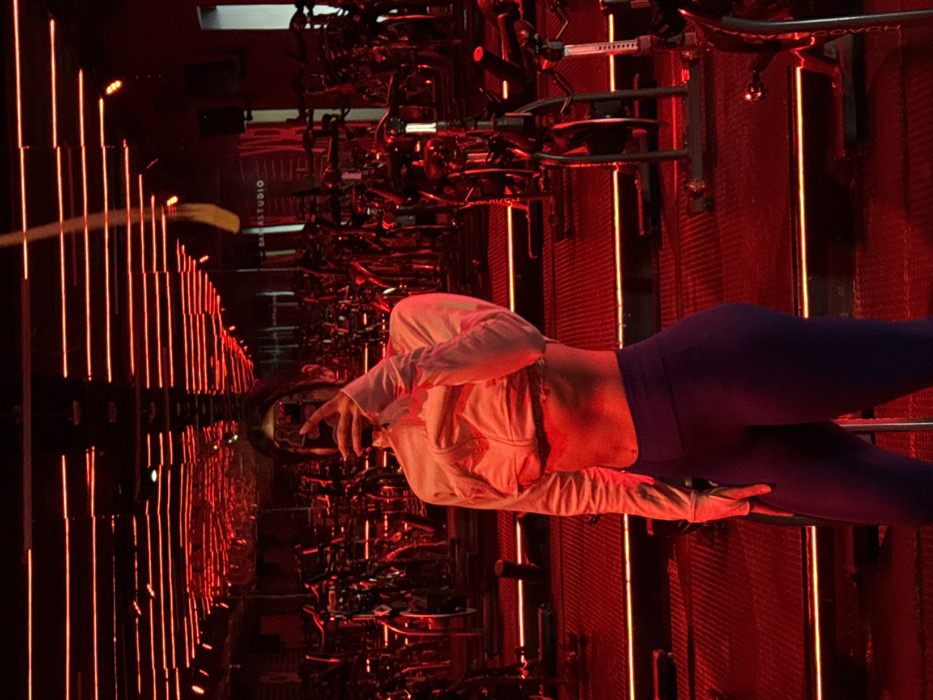 The image displays an individual taking a mirror selfie inside an indoor cycling or spin studio. The environment is bathed in a predominant red ambient light, originating from numerous horizontal linear LED strips on the ceiling, which are extensively reflected on the floor and mirrored walls. Rows of black stationary exercise bikes with metallic accents are visible throughout the studio, filling the background. The individual, an apparent female, stands in the foreground, dressed in athletic wear consisting of a light-colored, long-sleeved cropped top that exposes her midriff, and dark purple, form-fitting leggings. She holds a dark-cased smartphone at face level to capture her reflection, thereby obscuring her facial features. A partial sign in the background reflection clearly displays the text "BARRASTUDIO," identifying the facility. A yellow strap or rope is also faintly reflected, suspended from the ceiling. This studio is located within the metropolitan area of Puebla City, Mexico, specifically a BarreStudio facility, likely situated in the Reserva Territorial Atlixcáyotl district of San Andrés Cholula.
