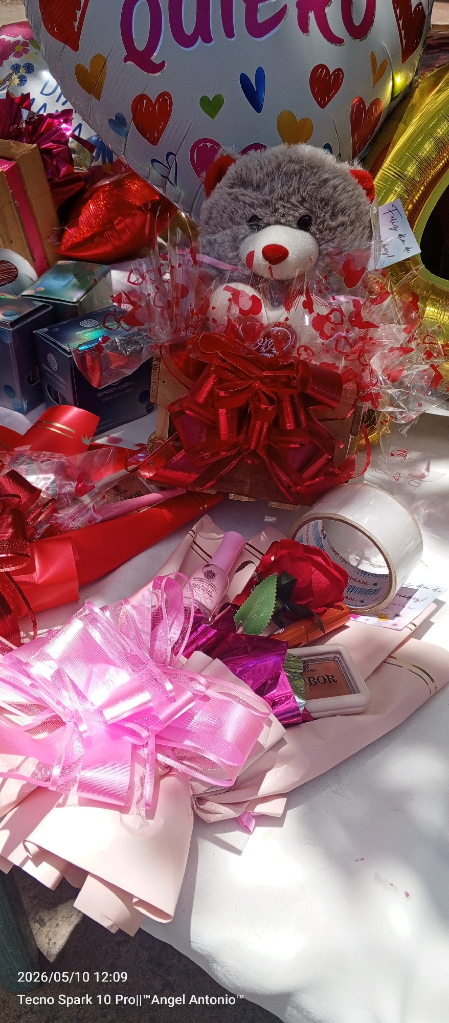 This image depicts a collection of gifts arranged on a surface, likely a table. The gifts include a teddy bear in a wooden basket adorned with red ribbons, wrapped boxes of cosmetic products (possibly from the brand "Grenobil"), a rose, pink nail polish, makeup (possibly "Nobor" brand), a roll of clear tape, pink and red ribbons, and balloons. The balloons include a large heart-shaped one with the Spanish word "Quiero" (I want/love), along with other decorated heart-shaped and gold balloons. A small handwritten note is attached to the teddy bear, likely a gift tag. The scene seems to be a celebration or gift-giving occasion. The location, Juan Griego, Venezuela, is not explicitly depicted. The information at the bottom of the image, "Tecno Spark 10 Pro||™ Angel Antonio™ 2026/05/10 12:09," indicates the photo was taken with a Tecno Spark 10 Pro mobile phone on May 10, 2026, at 12:09, by someone named Angel Antonio.