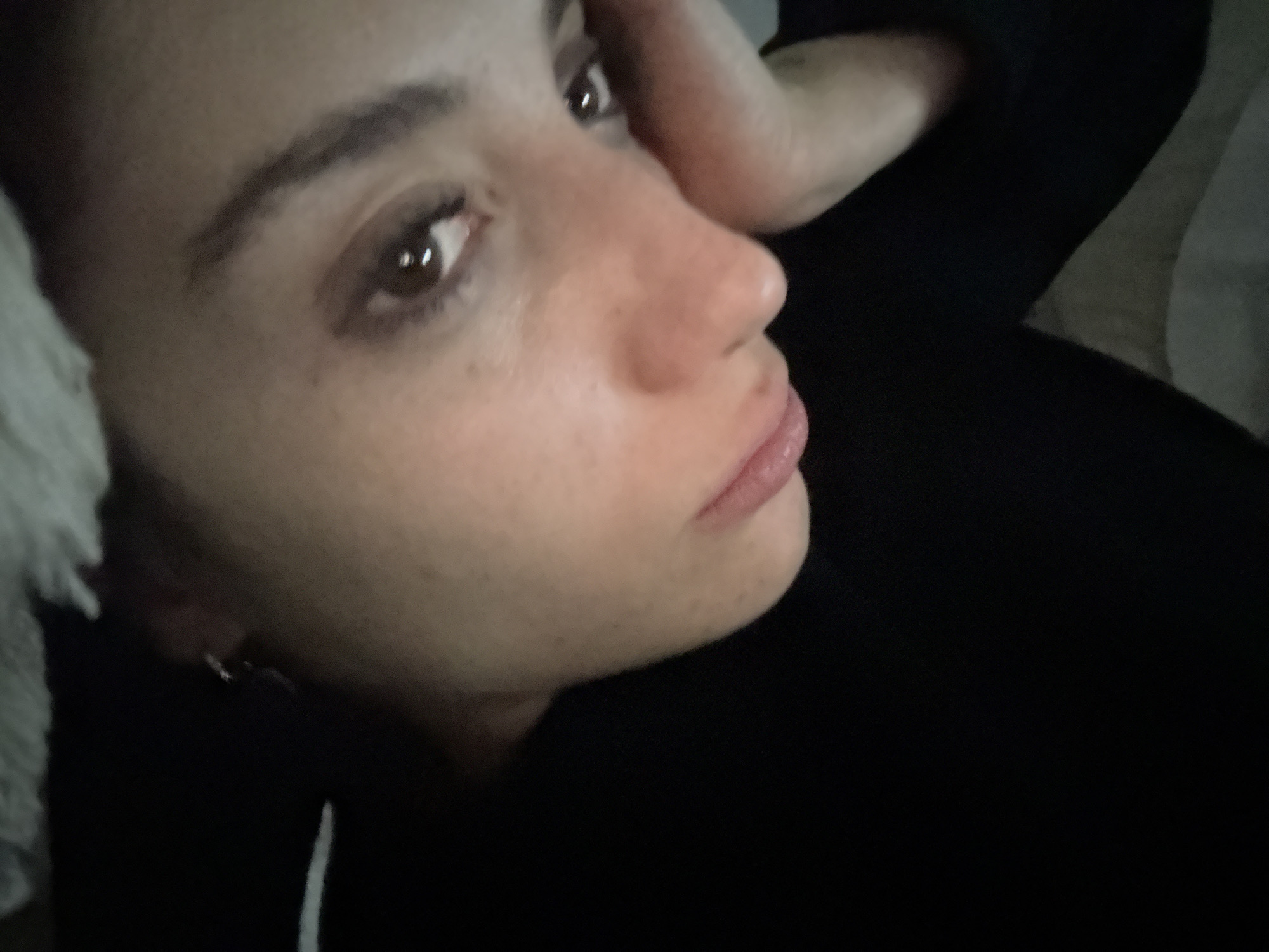 A close-up, low-angle shot captures a woman's face. Her head is tilted back, with her right hand near her head. She has brown eyes, dark eyebrows, and appears to be wearing a dark hoodie or shirt. A white object, likely a pillow, is visible to the left of her face. No information about the city of Buenos Aires, Argentina, is present.