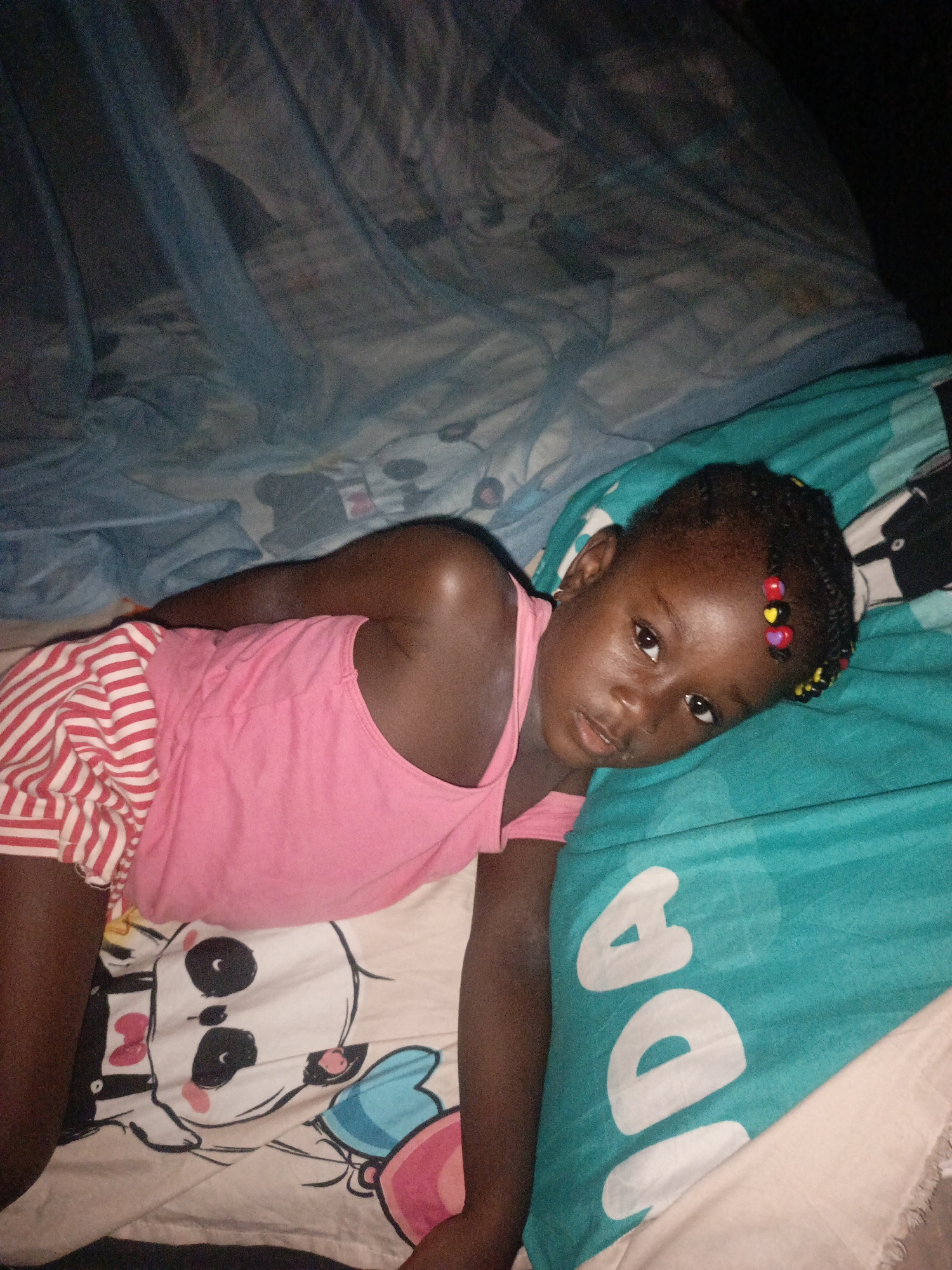 A young Black girl with braided hair adorned with colorful beads lies on a bed with patterned sheets and a turquoise blanket. She is wearing a pink tank top and red and white striped shorts. Her head is propped on her arm, and she is looking towards the camera. The room appears to be dimly lit, possibly at night. The bed is covered with a light blue mosquito net. The overall scene suggests a relaxed or sleepy atmosphere within a residential setting in Akwanga, Nigeria.