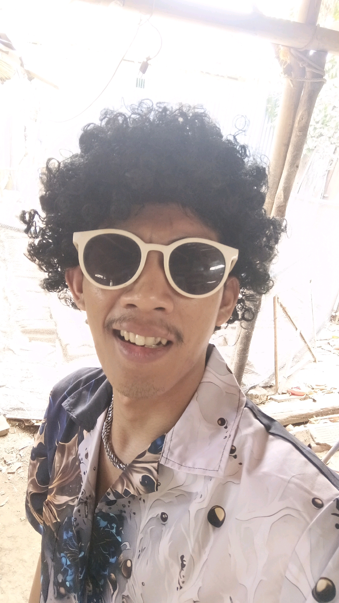 A person wearing a curly black wig, light-rimmed sunglasses, and a short-sleeved button-up shirt with a colorful abstract pattern is posing for a selfie. The shirt is predominantly white with swirls of dark blue, black, and tan, and features circular patterns with dark centers. A thick silver chain necklace is visible around the person's neck. The background appears to be an outdoor, somewhat rustic setting with wooden poles and beams, possibly suggesting a construction or unfinished area. The ground is covered with debris and loose soil. The overall lighting is bright, indicating daytime. The location is stated as Kyereku, Nigeria, however, this information is not visually verifiable from the image itself.