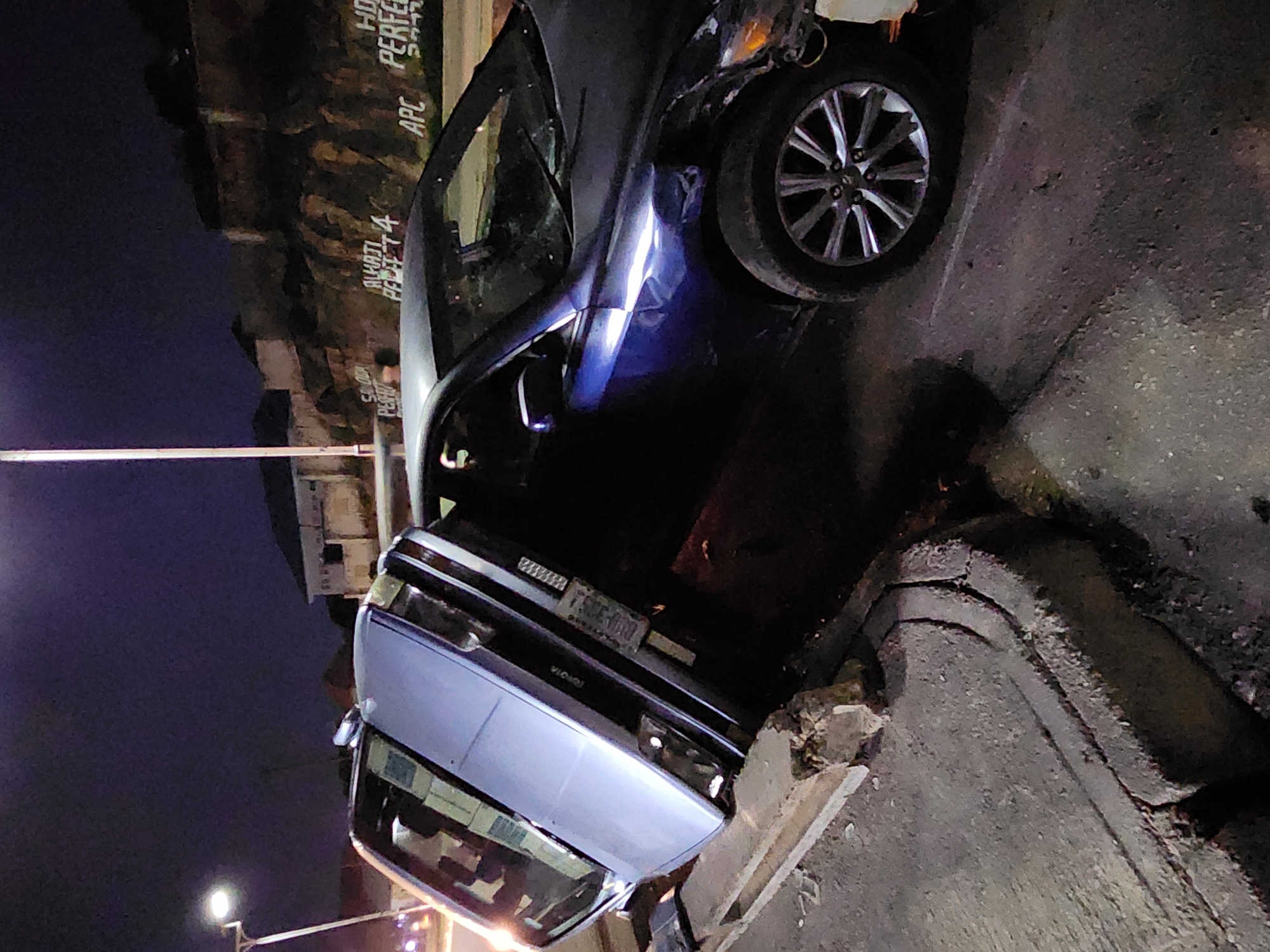A night scene shows a car crashed on a curb. The car, a dark blue sedan, is partially on the sidewalk, with its front end damaged. The driver's side door is open. The location appears to be an urban area, with buildings and signs visible in the background.