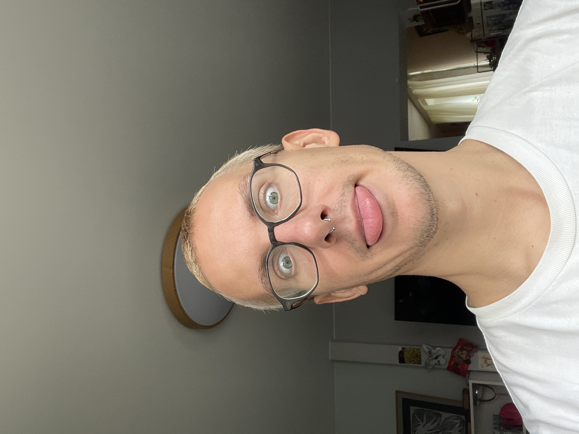 A close-up, portrait-oriented shot shows a person with short, bleached blonde hair wearing round glasses and a white t-shirt. The person has a fair complexion, a nose piercing, and is sticking out their tongue. The background includes a gray wall and a circular wall-mounted object with a light interior. Behind the person, other objects and furniture are visible, although less distinct. The location in the city of Sestroretsk, Russia cannot be determined from the visual information.