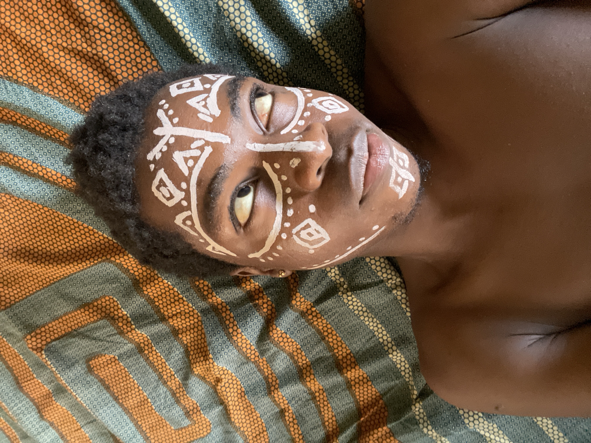A brown-skinned male with short, dark hair lies on a patterned fabric. His face is adorned with white painted tribal markings. He has dark eyes, and his lips are slightly parted. The fabric beneath him has a geometric pattern in shades of orange and green. The background is out of focus, with no specific indication of Nkpor, Nigeria.