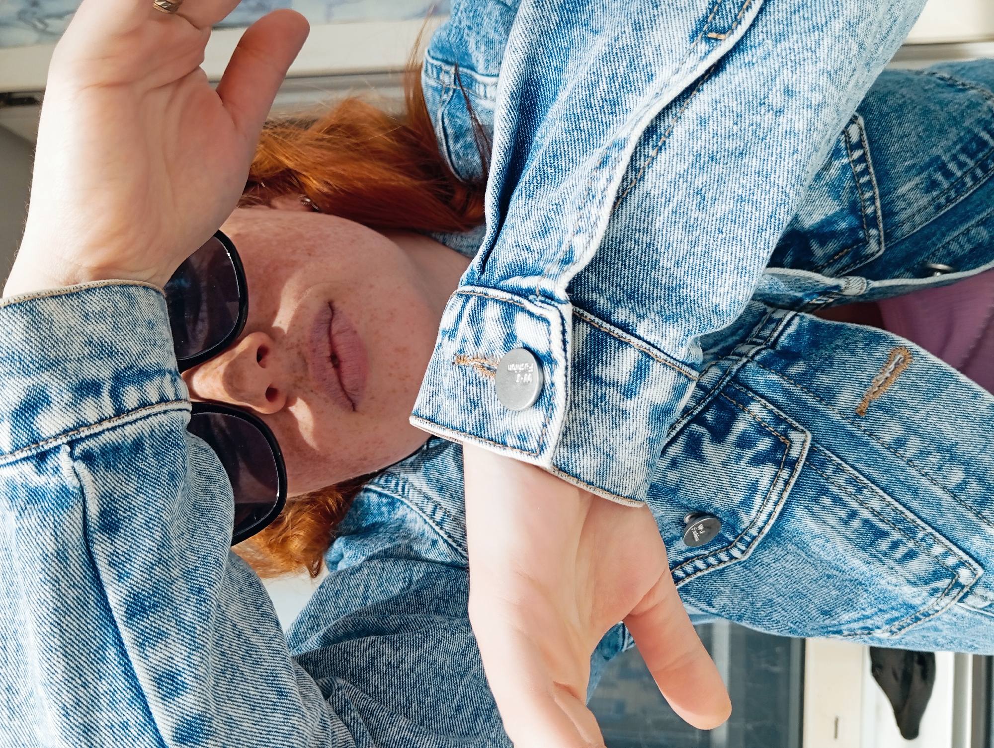 A red-haired woman with freckles wears sunglasses and a denim jacket. She is indoors, near a window. Her hands are raised in a pose. The city of Yurgamysh, Russia, is not explicitly visible or deducible from the image.