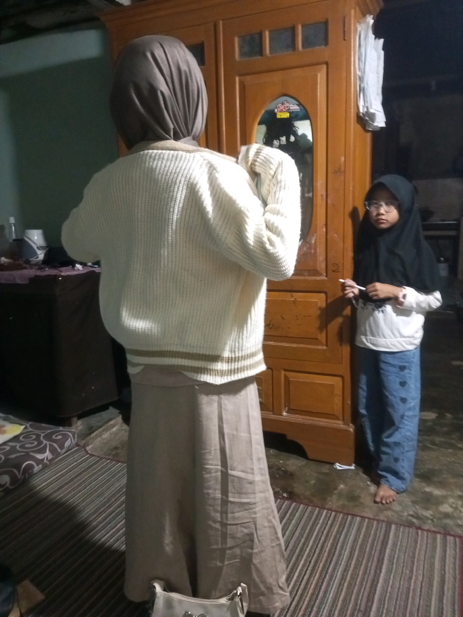 The image depicts an indoor scene, likely a residential interior, within the city of Andongsili, Indonesia. The primary subjects are two individuals, both appearing to be females, dressed in modest attire that includes headscarves. The individual in the foreground, facing away from the camera, is wearing a light-colored, textured sweater over a long, tan skirt. The second individual, positioned to the right and facing the camera, is wearing a black headscarf and glasses, a white long-sleeved shirt, and blue pants adorned with heart patterns. She is holding a white pen or stylus.
The environment includes a wooden wardrobe or cabinet with multiple doors and glass panels, reflecting a distorted image of another person. The flooring is a combination of what appears to be a patterned rug and a tiled or concrete surface. A dark-colored piece of furniture, possibly a dresser or cabinet, is visible on the left side of the frame. The lighting suggests it might be evening or a dimly lit interior. There are no obvious indicators of specific urban landmarks of Andongsili.