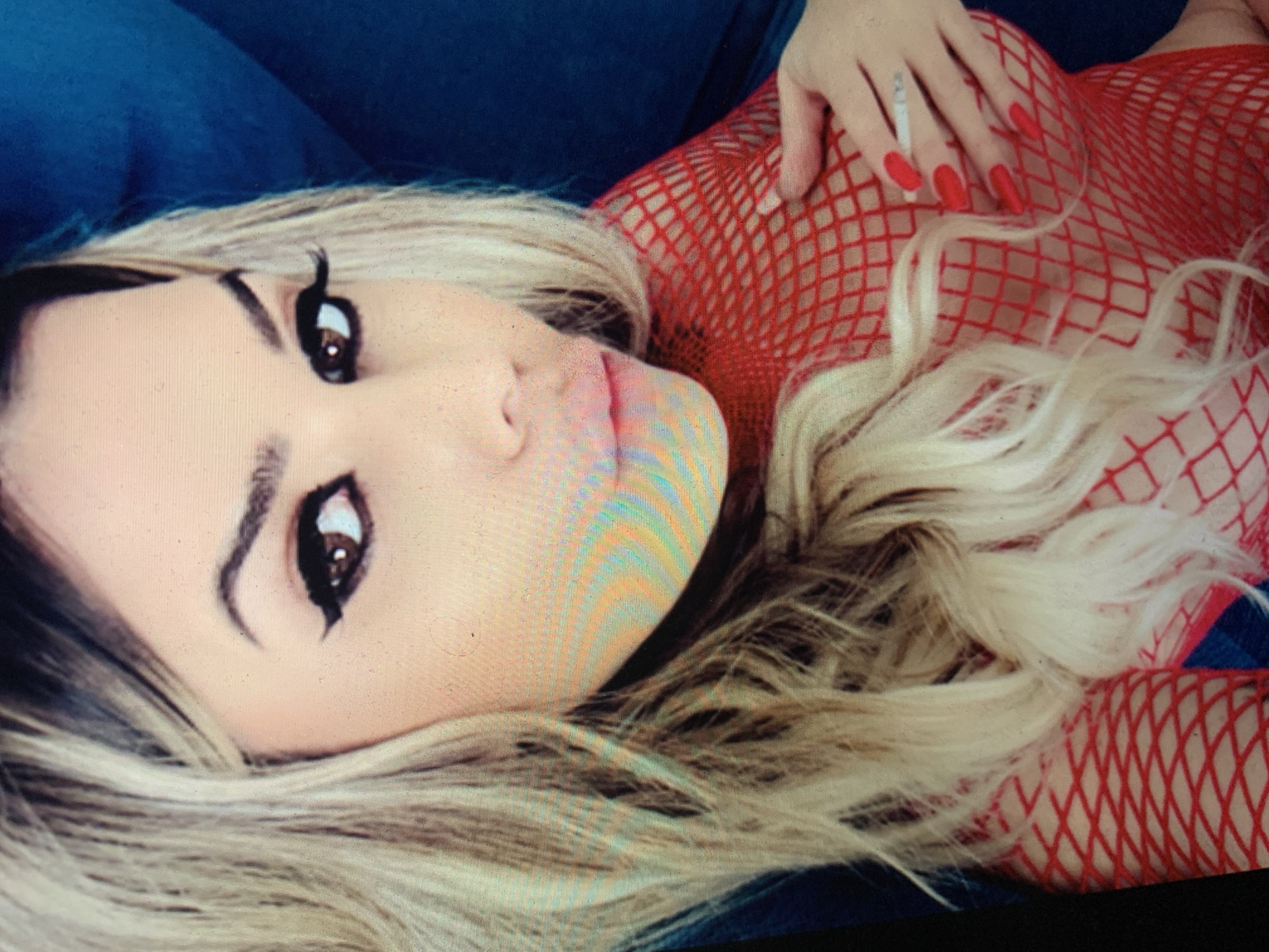 An individual with blonde hair and heavy makeup, including prominent winged eyeliner and bold lipstick, is captured in a close-up portrait. They are wearing a red fishnet top and have bright red fingernails. One hand is placed on their chest, and the other holds a lit cigarette between two fingers. The background is dark and out of focus, with a hint of a blue surface visible on the right. The overall lighting suggests an indoor setting.