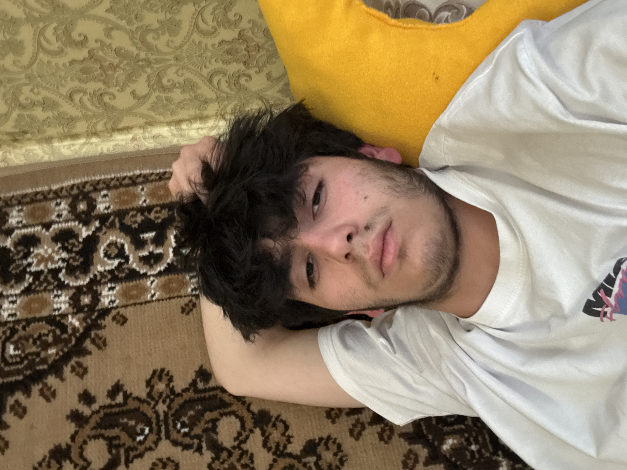 A young man with dark hair and a beard is lying down, facing the camera. He is wearing a white t-shirt and resting his head on his hand. A yellow pillow is behind his head. He is lying on a patterned carpet next to a patterned wall. The location cannot be determined as Yerevan, Armenia, based on the image alone.
