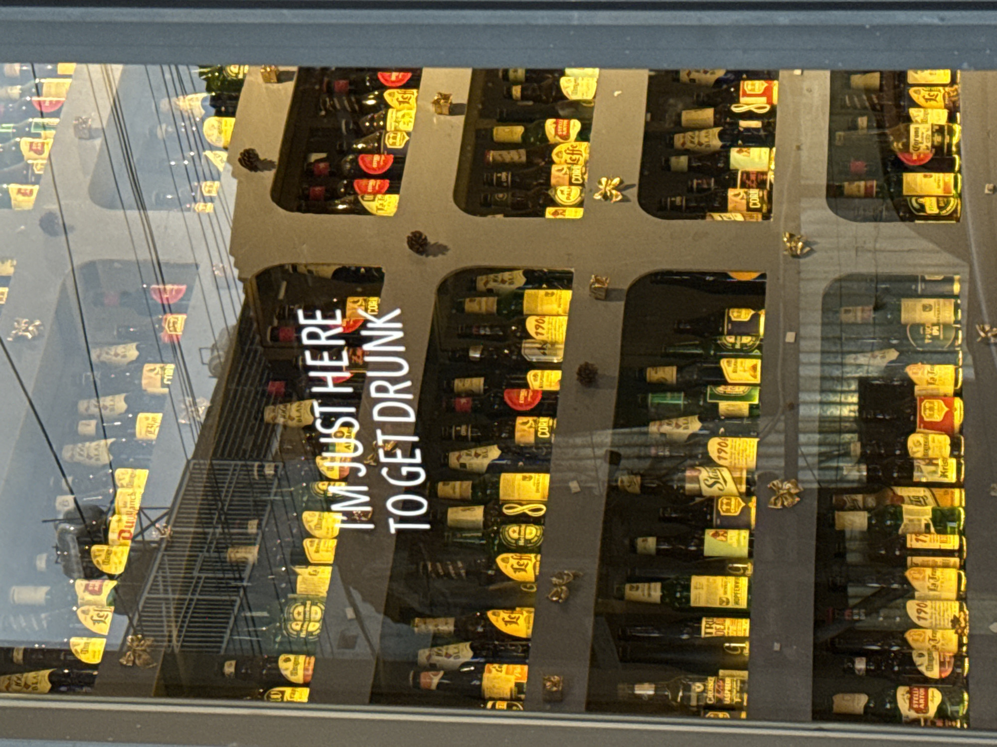 The image captures an illuminated display of numerous alcohol bottles, predominantly beer and wine, showcased on shelves behind a glass window. The bottles are arranged horizontally within individual compartments, creating a grid-like visual. Many labels are brightly colored in yellow, red, and white, contributing to a warm glow emanating from the display. Small decorative items, including golden bows and pinecones, are placed intermittently among the bottles.
A clear reflection on the glass surface overlays the bottles, revealing elements of the surrounding environment, such as a multi-level structure with railings, hinting at a building across the street or another part of an interior space.
A prominent, vertically oriented text is superimposed on the glass, stating "I'M JUST HERE TO GET DRUNK" in large white letters. This phrase serves as a humorous and direct message.
The artificial lighting suggests an indoor commercial setting, likely a liquor store or bar in Da Lat, Vietnam. The overall presentation is vibrant and well-stocked.
Visible text on various bottle labels includes "BLA" (Blanc), "CRUISI", "RASPBERR", "TRU", "HOPFENW", "Cuollanche Blanc", "COR" (Corona), "190", "PALH", "Coffe", "Stay 1904", "SILAN", "STELLA ARTOR" (Stella Artois), "La Tra", "Krist", and "IPA", indicating a diverse selection of beverages.