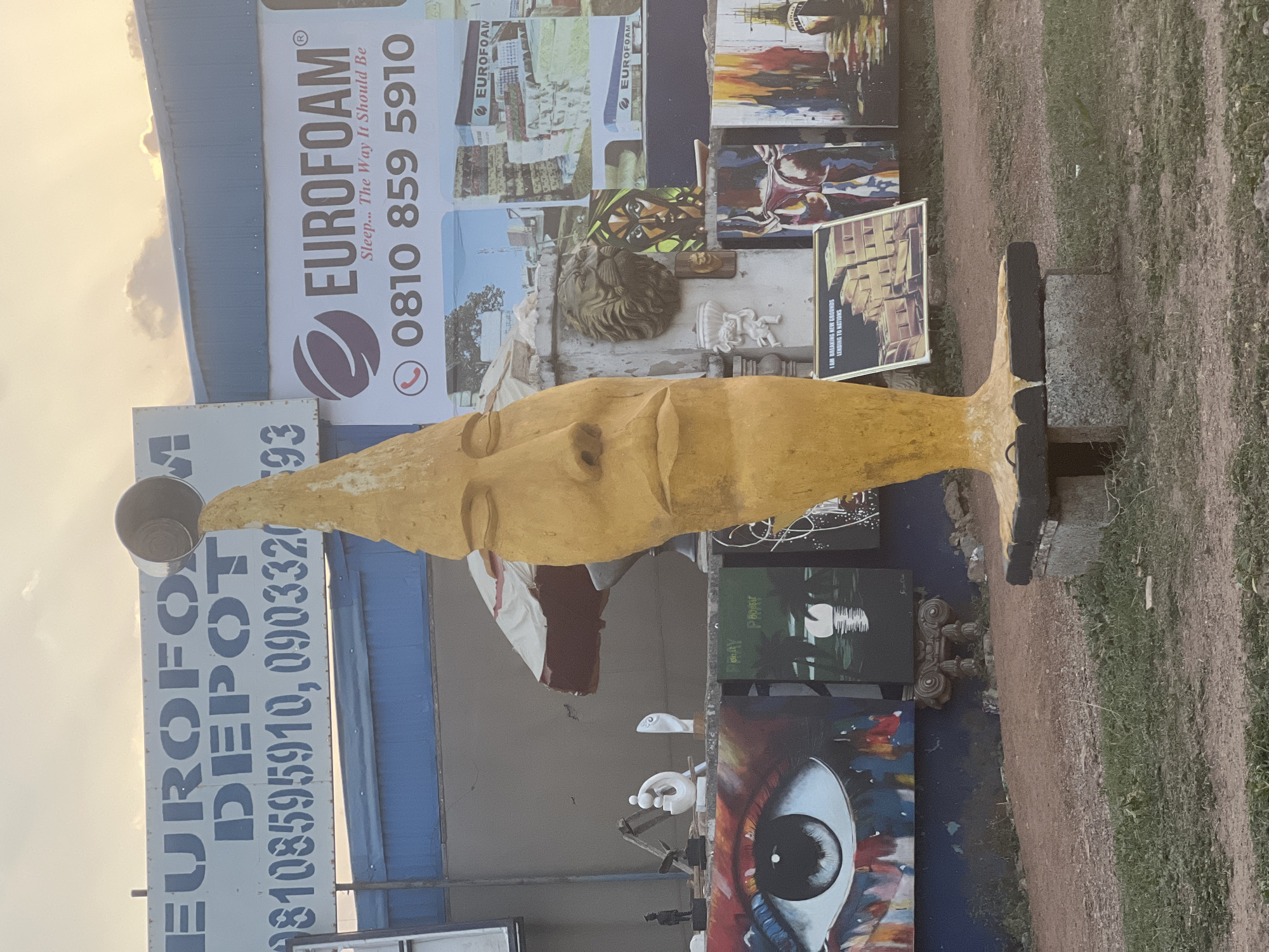 The image depicts an outdoor display of artwork and sculptures in front of a building with "EUROFOAM DEPOT" and "EUROFOAM" signage, along a dirt path. The location is likely in Abuja, Nigeria, given the phone numbers on the signs. The main subject is a large, elongated yellow sculpture of a face. Behind it, various paintings and smaller sculptures are arranged, including artwork featuring a large eye and others with abstract designs. The background includes commercial signage with phone numbers. The ground is a mix of dirt and grass. There are no people visible in the immediate foreground.