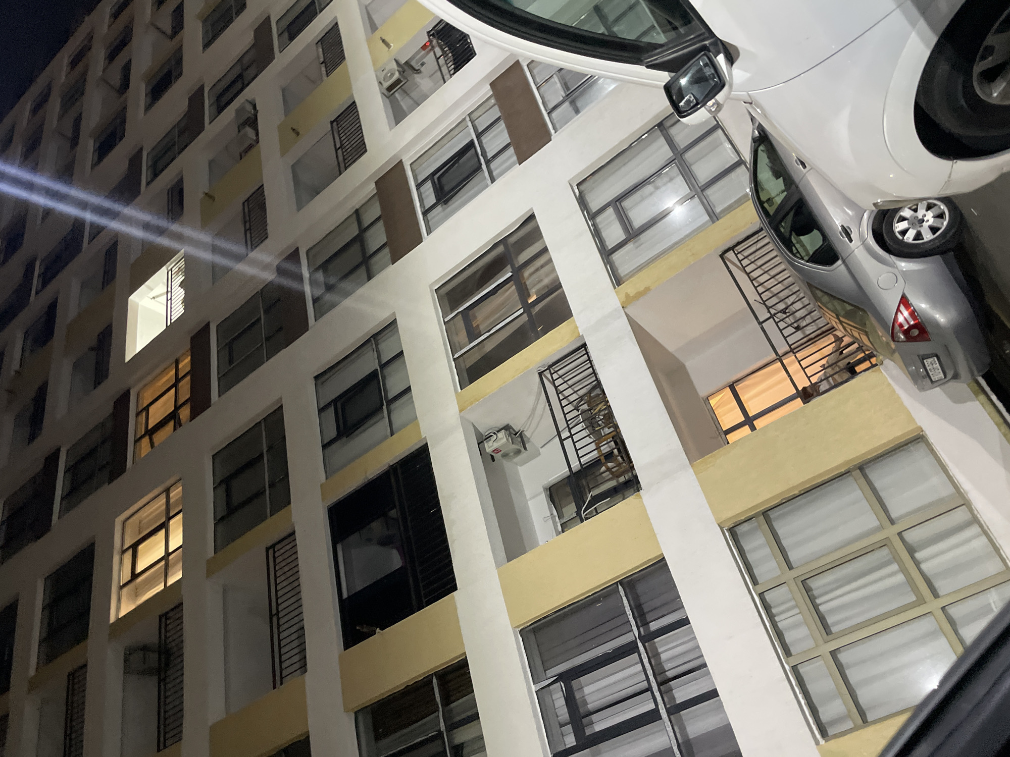 The image shows a nighttime view of a residential building in Sumgayit, Azerbaijan. The building is tall and appears to be made of concrete with yellow and white accents. Many windows are visible, some illuminated from within, suggesting occupancy. Balconies with black railings are present on several floors. In the foreground, the rear of a silver sedan is parked next to a white vehicle, which is partially visible on the right side of the frame. The lighting suggests it is after dark, with artificial lights from inside the building and potentially streetlights illuminating the scene. The overall impression is of an urban residential environment.