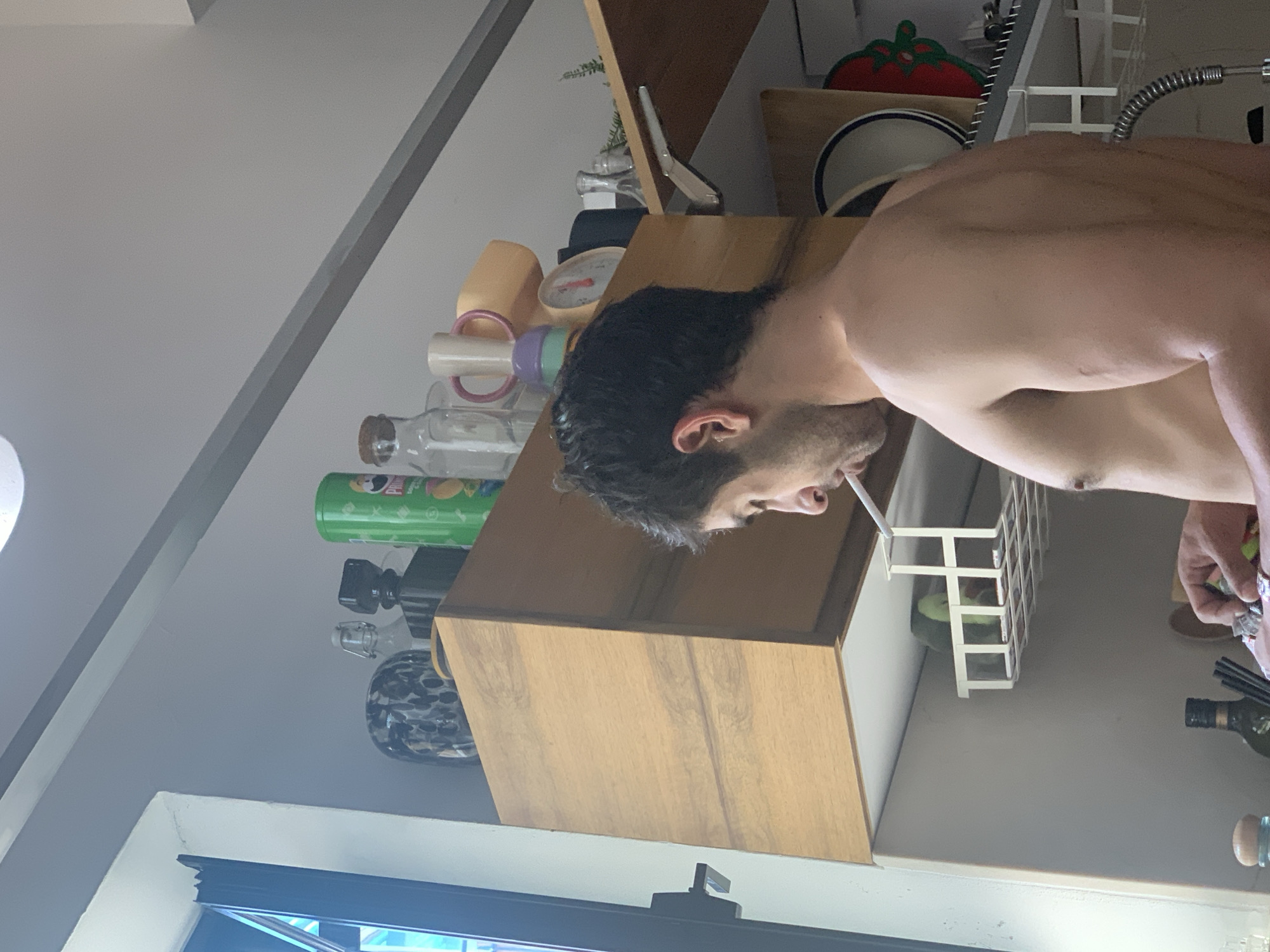 The image captures an indoor scene, likely a kitchen or living area in a home, with a man standing in profile on the right side of the frame. The perspective is rotated, showing the man and the counter he is at from above, with the room's ceiling on the left.
The main subject is a man, visible from the chest up. He has dark, wavy hair and stubble on his face. He appears shirtless or is wearing a light undershirt not fully visible. A cigarette or similar item is held between his lips. His expression is neutral, and he seems focused on an activity with his right hand, which is partially obscured, possibly engaged in preparing something or holding an item near the counter.
The background reveals a cluttered but organized counter space. On a light wooden cabinet or shelf, various items are placed: several glass bottles (one with a cork, another with a patterned design), a green Pringles can with the logo and a cartoon character visible, a yellow cup with a pink handle, and a small scale with a red needle. Further back on the counter, there's a silver kitchen utensil, a small red tomato-shaped object (possibly decor or a timer), and a white bowl with blue stripes. A white dish rack/drainer holds some green items, and a flexible metallic kitchen faucet is visible above it. Towards the lower right, a dark bottle (possibly olive oil) and another clear bottle are present. A bright light source is partially visible on the far left. A window frame is visible in the bottom left corner, suggesting natural light entering the space.
The overall impression is a candid shot of someone in their home, possibly taking a break or involved in a casual kitchen activity. The time of day appears to be daytime due to the natural light. No explicit visual cues in the image confirm the Tel-Aviv, Israel location, but the setting is typical of a residential interior.
