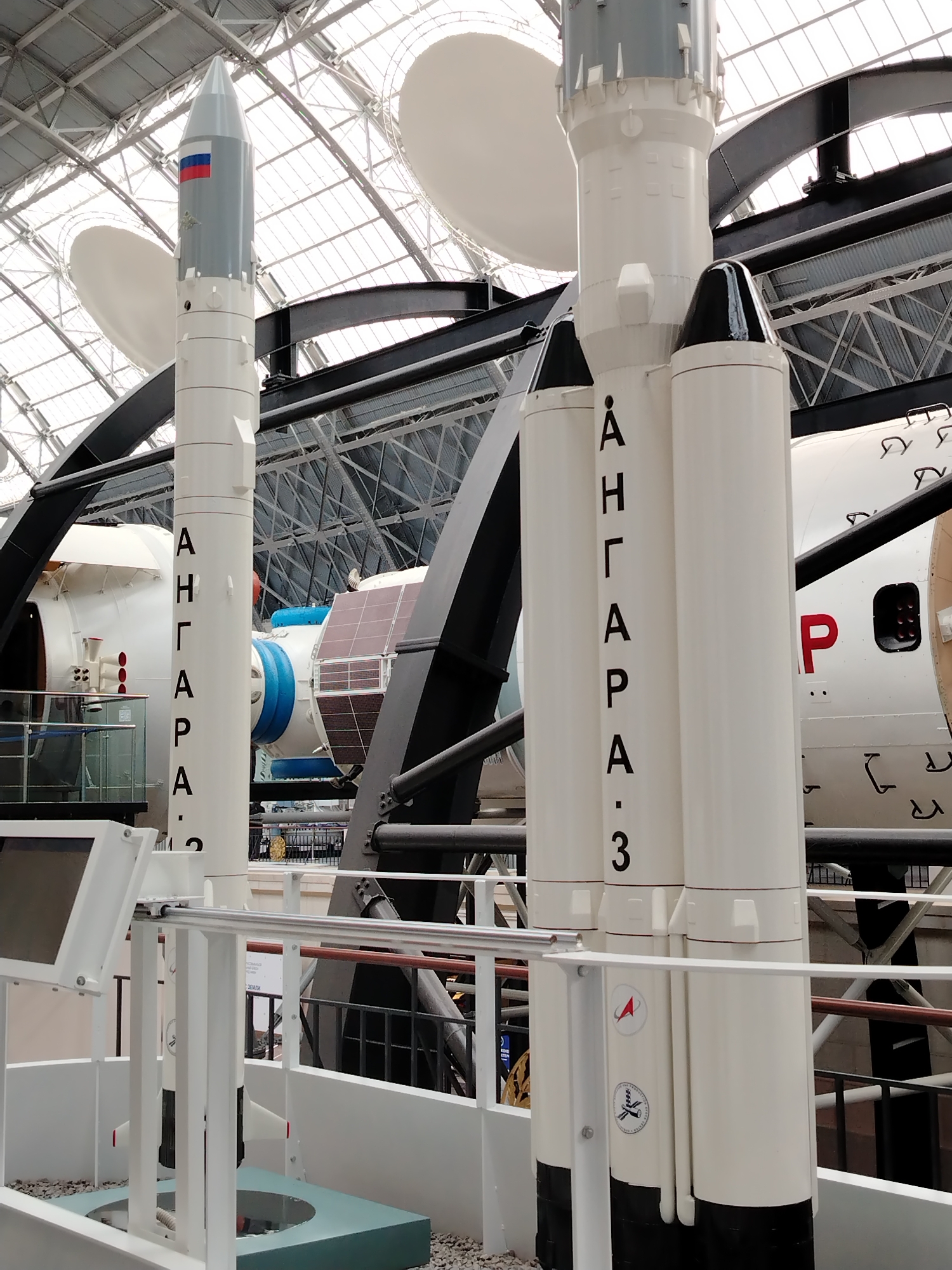 This is a medium shot of a display of rockets and space artifacts inside a large, modern building with a glass and metal roof structure. In the foreground, a white rocket stands vertically, with black lettering that reads "АНГАРA.3" vertically down its side. To the right of this rocket are two other white rocket segments, one slightly taller than the other. Both have black conical tips. The rocket on the left has a circular emblem with a red arrow. The rocket on the right has another circular emblem with a laurel wreath and a symbol of a rocket launching. Behind the rockets is a large white cylindrical object with solar panels and a red emblem that appears to be the Soviet Union's hammer and sickle. A window or hatch is visible on the side of this object. The background consists of the building's interior structure, including metal beams and the translucent ceiling. The lighting suggests an indoor, well-lit environment. This display is likely part of a museum, potentially the Cosmonautics Museum in Moscow, Russia, given the Russian text and space-related theme. There are no people visible in the image.