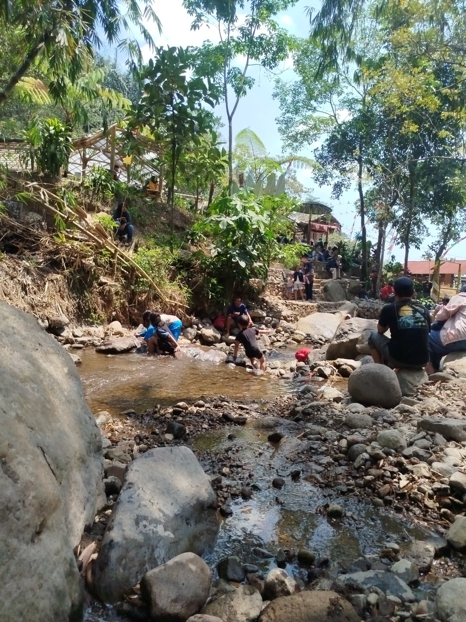 The image depicts a natural environment, likely a river or stream flowing through a rocky terrain. Lush green foliage and trees dominate the background, with a clear blue sky visible through the canopy. In the foreground, several large rocks line the banks of the stream. A group of people are present, some of whom are wading in the shallow water. Others are seated on the rocks or on a raised platform further back, suggesting a recreational area. The presence of a simple, open-air structure with a thatched roof and flag decorations indicates a possible rest area or gathering spot. The overall scene suggests a peaceful outdoor gathering in a natural setting, possibly in the region of Ngesrepbalong, Indonesia, known for its natural landscapes.