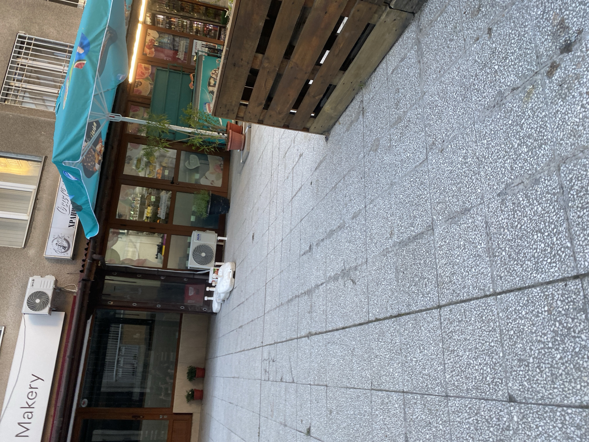 The image shows a street in Sofia, Bulgaria. In the foreground, there is a grey tiled sidewalk. Adjacent to the sidewalk, a wooden pallet is propped up against the wall of a building. The building houses a store named "Makery," as indicated by the sign on the storefront. The storefront features glass windows and doors. An air conditioning unit is mounted on the building's exterior. A turquoise umbrella is positioned in front of the store. Additional air conditioning units are visible above the storefront. There are flowerpots outside the door.