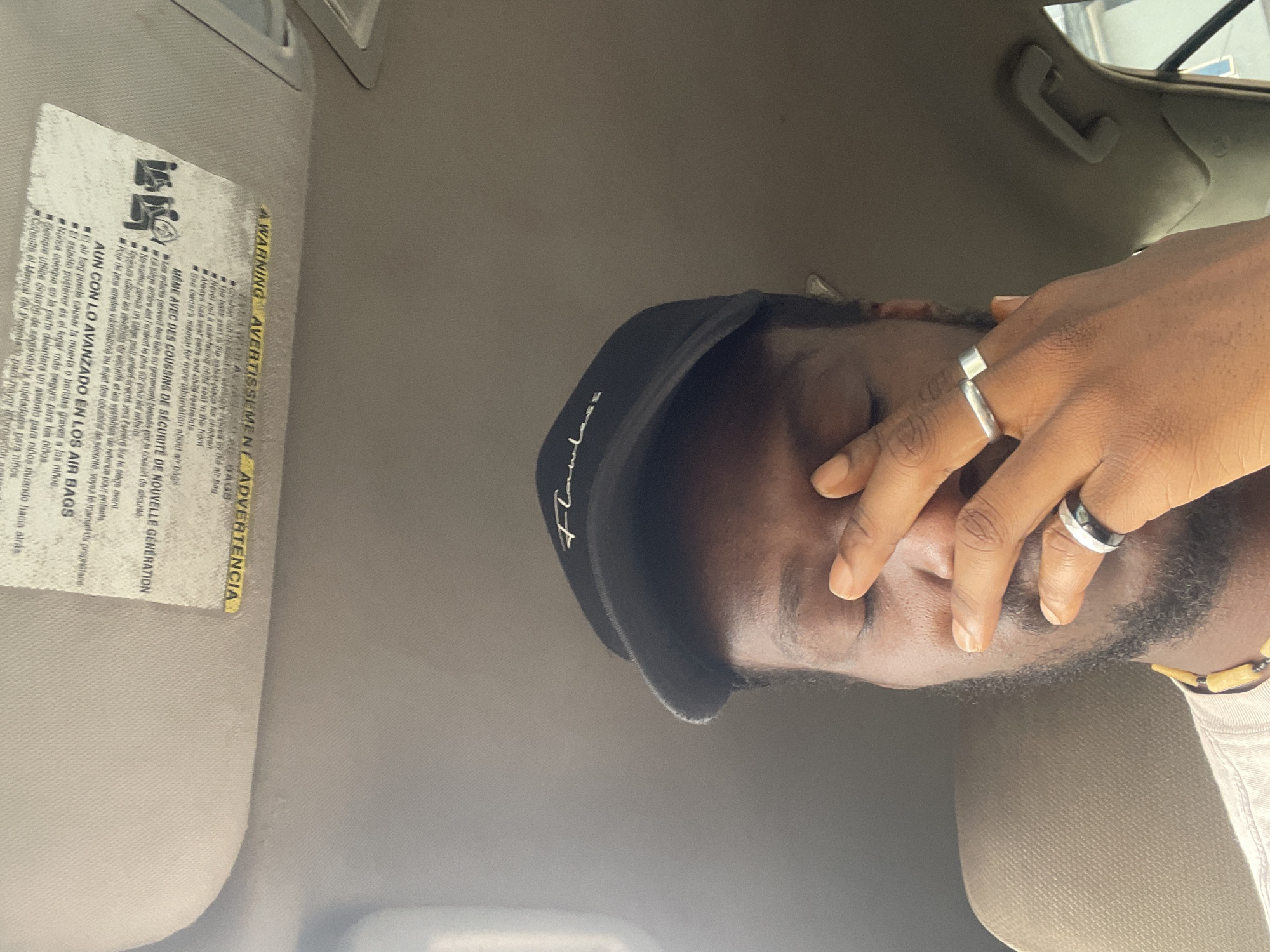 This media file shows a close-up, rotated shot of a person inside a vehicle, likely a car, as indicated by the sun visor with warning labels. The person is wearing a black baseball cap that says "Flawless" in white script. Their right hand is held up, covering their face, with two rings on their fingers. One ring is a simple silver band, and the other appears to be a darker band with a silver inlay.
The sun visor displays warning labels in English, French, and Spanish regarding airbags and child safety. The text includes phrases like "WARNING," "AVERTISSEMENT," and "ADVERTENCIA," along with instructions to never place a rear-facing child seat in the front seat and that the back seat is the safest place for children.
The lighting suggests it might be daytime, possibly with natural light filtering into the car. The background is blurred and appears to be the interior of the vehicle, with a handle visible on the ceiling. The person has dark skin and a beard, and their eyes are not visible due to their hand covering their face. The overall mood is somewhat obscured or contemplative, with the hand hiding the expression. The rotation of the image makes it appear as if the viewer is looking at the scene from a sideways perspective.