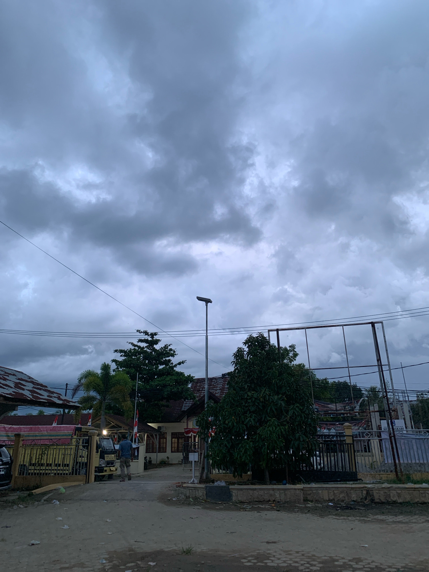This image depicts an outdoor scene in Pantegurah, Indonesia, under a heavily overcast sky with dark, textured clouds filling the upper portion of the frame. The lighting suggests it is either early morning or late afternoon.
In the foreground, a dirt and gravel road leads into a property. To the left, a gate with a yellow and red striped truck parked behind it is visible. A man wearing a blue shirt and camouflage pants is walking away from the camera towards the truck and a building. The building appears to be a single-story structure with a brown-tiled roof and visible windows. A palm tree stands to the left of this building, and another leafy tree is situated to the right of the building.
Further to the right, a large, empty metal frame structure stands, possibly a billboard or sports equipment frame. Behind this frame, another building with a tiled roof is partially visible, along with fencing and what appears to be banners or signs. Electrical wires are strung across the scene, and a lamppost is positioned in the middle ground. The overall impression is a somewhat utilitarian area, possibly residential or commercial, with evidence of infrastructure like roads, buildings, and utilities.
