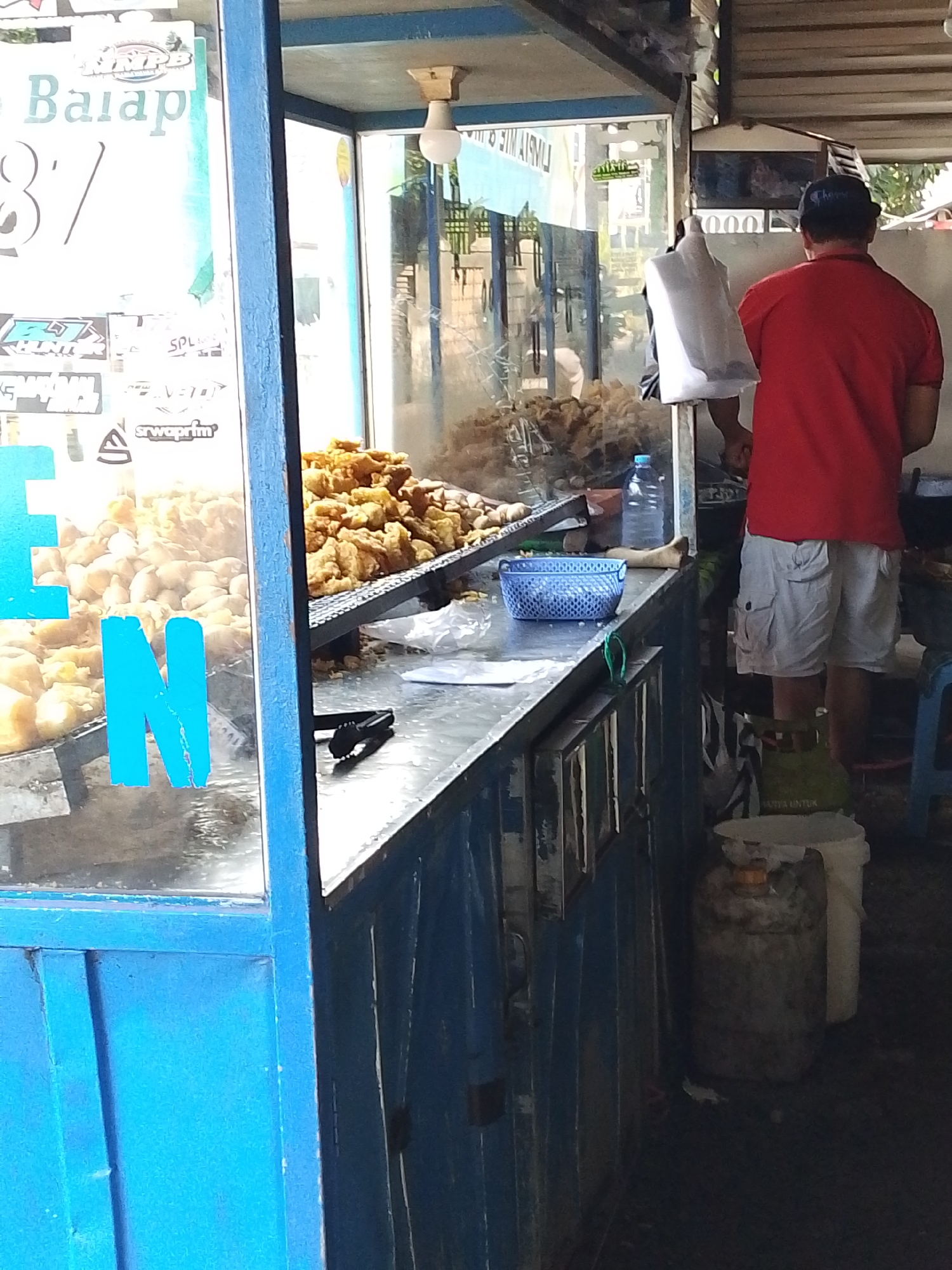 In Kediri City, Indonesia, a street food vendor operates a stall selling fried food, possibly chicken or fish. A man in a red t-shirt and white shorts is standing behind a metal counter, facing away from the camera, and appears to be attending to the food. He is holding a white plastic bag. In front of him, a blue woven basket sits on the counter next to a bottle of water and a pair of black tongs. Piles of golden-brown fried food are displayed on a metal rack behind the counter. The stall is characterized by its blue metal structure and a clear glass enclosure. Various stickers are visible on the glass, some featuring text and logos, and two large blue letters, "E" and "N", are affixed to the glass in the foreground. The environment is dimly lit, suggesting it may be an outdoor stall under a roofed structure. Two gas cylinders are visible on the ground to the right of the stall.