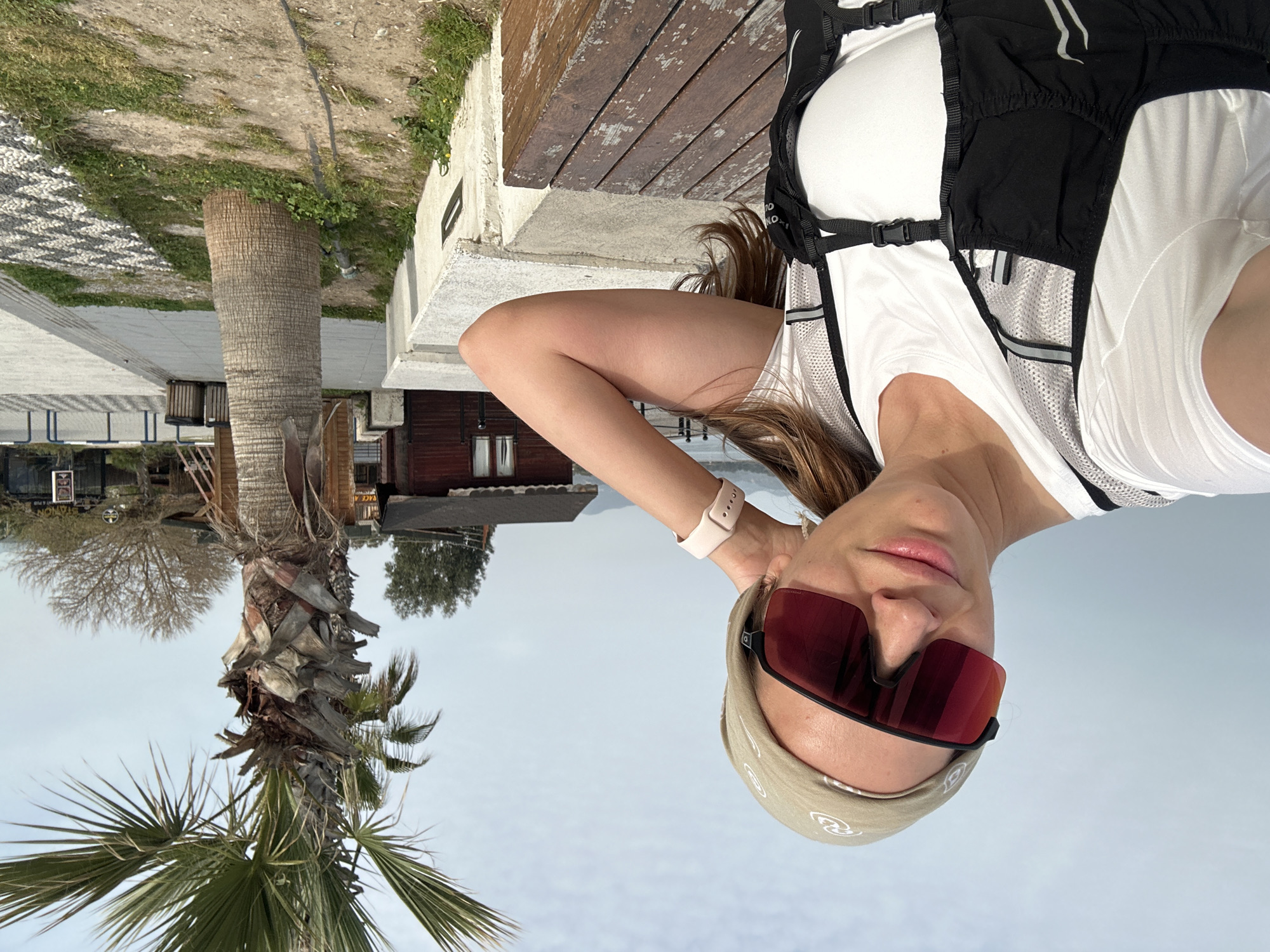 A woman wearing a white athletic top with a black hydration pack, a beige headband, and red sunglasses stands next to a palm tree. The location is Fethiye, Turkey, recognizable by the coastal architecture, pavement style, and wooden buildings in the background.