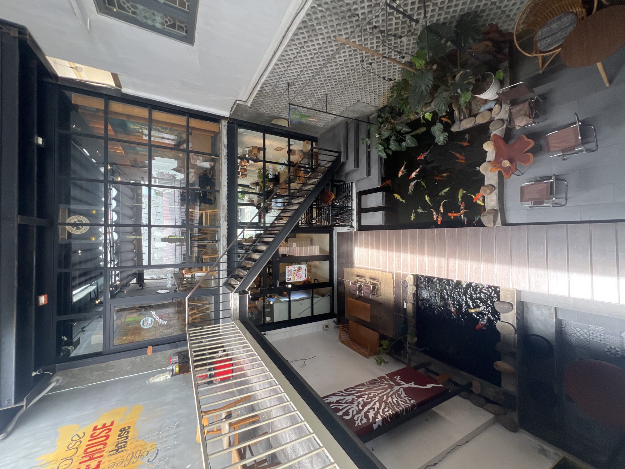 This is an interior shot looking down into a multi-level cafe in Hoi An, Vietnam. The cafe features an indoor koi pond with numerous fish, surrounded by lush greenery and decorative stones. A wooden walkway leads from the stairs to the pond area. On the right side of the pond, there are several chairs and a uniquely shaped wooden table with a drink on it.
On a lower level, to the left of the pond, there is a seating area with a wooden sofa and a coffee table. A large artwork of a white tree on a red background is displayed on the wall.
Above the pond and seating area, a staircase leads up to a mezzanine level, accessible from the ground floor. This mezzanine level is enclosed by glass walls and houses more seating areas and tables, with people visible dining or socializing.
The cafe has an industrial-chic aesthetic, with exposed metal beams and dark-colored stairs. The exterior walls are a mix of grey concrete and white painted surfaces, with some graffiti art visible on the left side of the upper wall, including "COFFEE HOUSE" in large, colorful lettering. The upper levels offer views of tables and chairs, with a balcony overlooking the ground floor and pond. Natural light streams in from the large windows, illuminating the space.