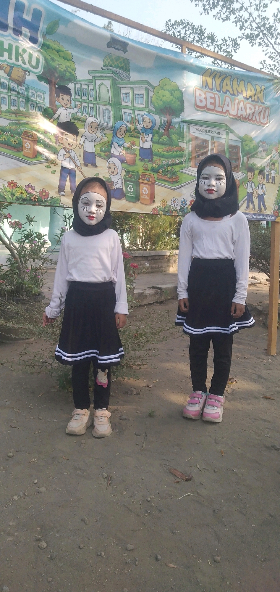 Two young girls, each wearing a black headscarf and white face paint, stand outdoors in front of a large banner. The banner depicts a colorful cartoon scene of children and a school building. The girl on the left wears a white long-sleeved shirt and a black skirt with white stripes at the hem, paired with black leggings and light-colored sneakers. The girl on the right wears a similar outfit: a white long-sleeved shirt, a black skirt with white stripes, and pink and white sneakers. The ground is dirt and gravel, with some dried leaves and twigs scattered about. In the background, there are plants and trees, and the sky is visible above. The banner has text that reads "NYAMAN BELAJARKU" and "POJOK KEBERSIHAN". This scene is located in Plemahan, Indonesia.