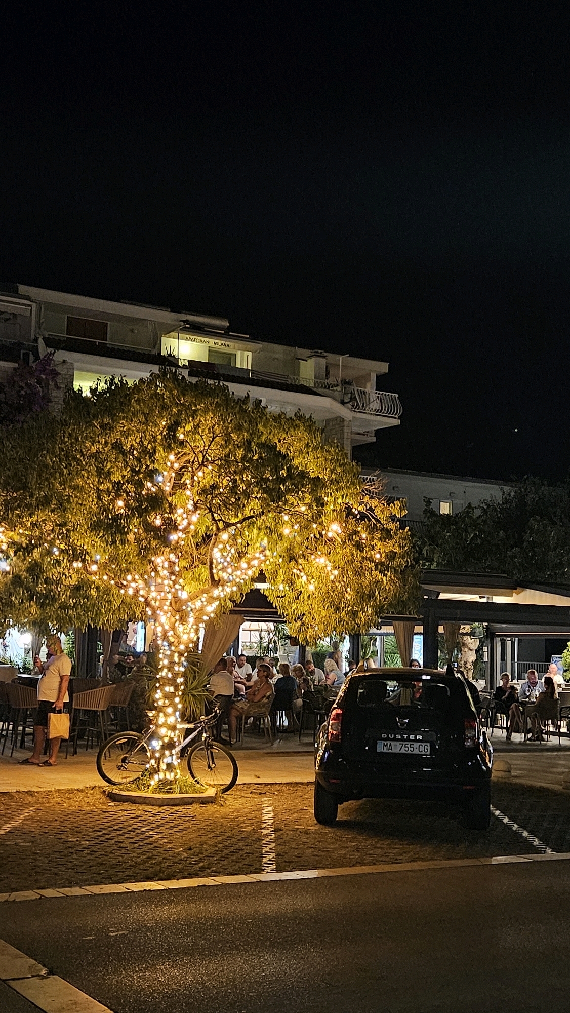 The image captures a nighttime scene in Općina Tučepi, Croatia, at what appears to be an outdoor dining area. A large tree, illuminated by numerous string lights, dominates the foreground, casting a warm glow over the surrounding space.
In front of the tree, a bicycle is parked, leaning against the tree trunk. To the right of the bicycle, a black Dacia Duster car is parked, its license plate reading "MA 755-CG".
Behind the tree, several people are seated at tables, suggesting a restaurant or cafe setting. Some patrons are visible in the background, engaged in conversation. A building, possibly apartments or a hotel, is situated behind the tree and dining area, with illuminated windows and balconies visible. The sky is dark, indicating it is nighttime. The ground is paved with interlocking stones.