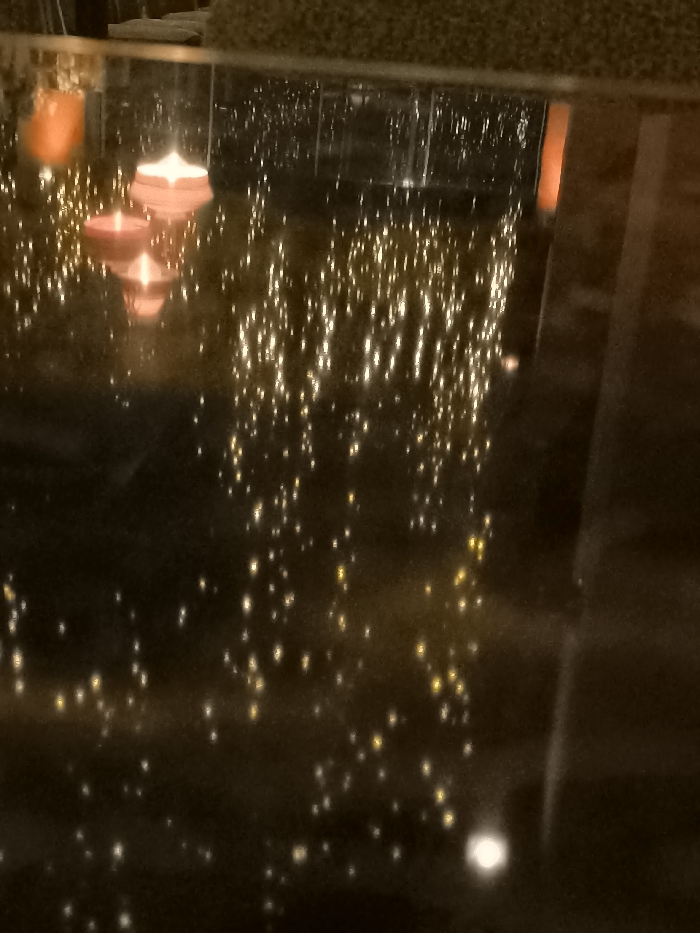 The image captures a serene nighttime scene featuring two floating candles on a dark, highly reflective surface, likely water. One candle is red and the other is pink, both emitting a soft glow, with their reflections clearly visible beneath them. The surface is adorned with countless small, bright, shimmering reflections, resembling scattered fairy lights or bokeh, creating a magical and ambient atmosphere. These lights appear to be reflections of decorative lights from the surrounding environment.
In the background, blurred vertical structures, possibly railings or window frames, and a fuzzy, textured object at the top (perhaps part of a sofa or upholstered seating) are dimly discernible. The overall impression is one of tranquility and romance, suggestive of a restaurant, hotel lounge, or a private residence in Palaio Faliro, Greece. The time of day is clearly night or very low light. No people or visible text are present.