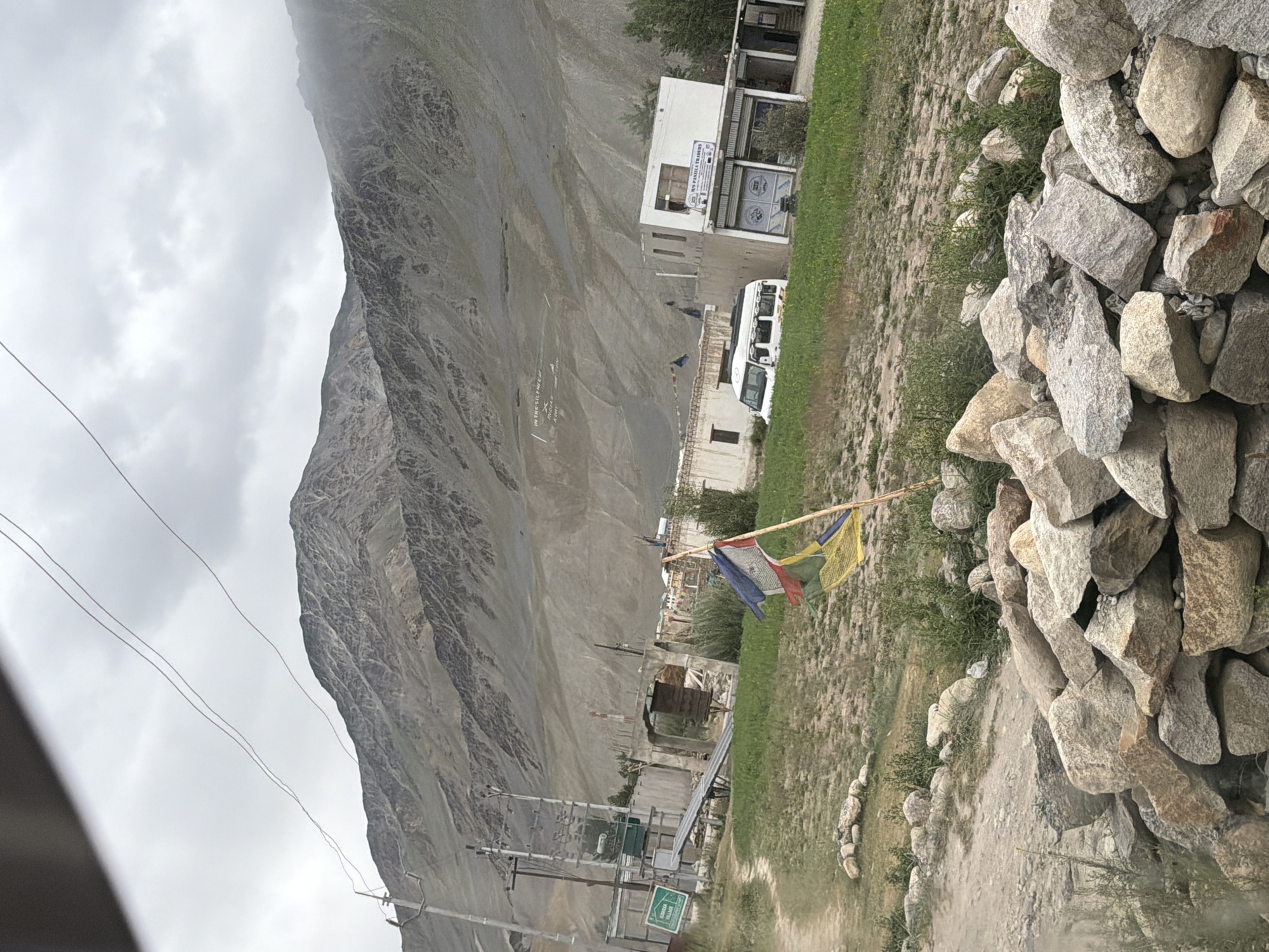 The image shows a view of Padum, India, with a mountainous backdrop under a cloudy sky. In the foreground, a pile of large rocks is visible. A dirt path winds through a grassy area. Several buildings are present, including a white building with signs and a garage-like entrance, and other structures that appear to be homes or shops. A white van is parked in front of one of the buildings. A prayer flag on a wooden pole is fluttering in the wind. There are also power lines and utility poles in the scene. On the side of the mountain in the distance, white lettering is visible. The overall impression is of a rural or semi-urban environment in a mountainous region.