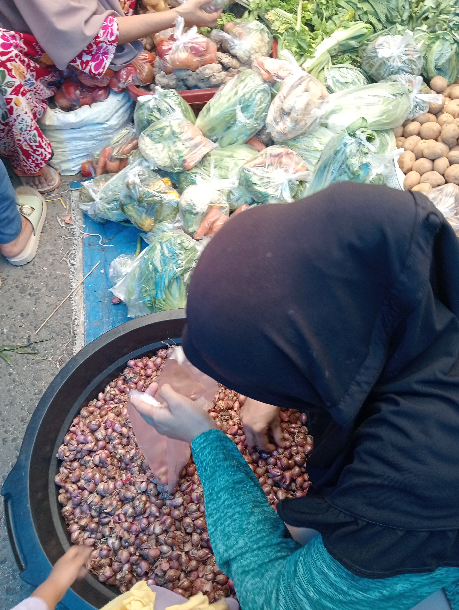This is a close-up, overhead shot of a market stall in Pariaman, Indonesia, focusing on a woman in a black hijab and teal top sorting shallots. The woman's hands are visible, one holding a plastic bag and the other sifting through the shallots in a large black tub. In the background, various vegetables are displayed in plastic bags, including what appear to be potatoes, cabbage, carrots, and tomatoes. The ground is paved and a blue tarp is visible. The overall scene suggests a bustling outdoor market.